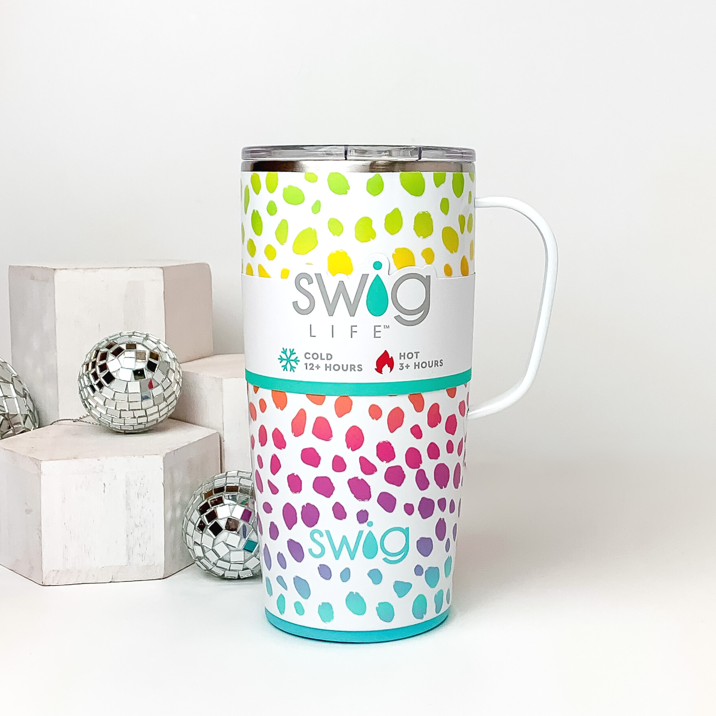 White mug with a clear lid and white handle. This mug includes a multicolored dotted print that is ombre. This mug is pictured on a white background with white blocks and disco balls on the left side of the picture. 