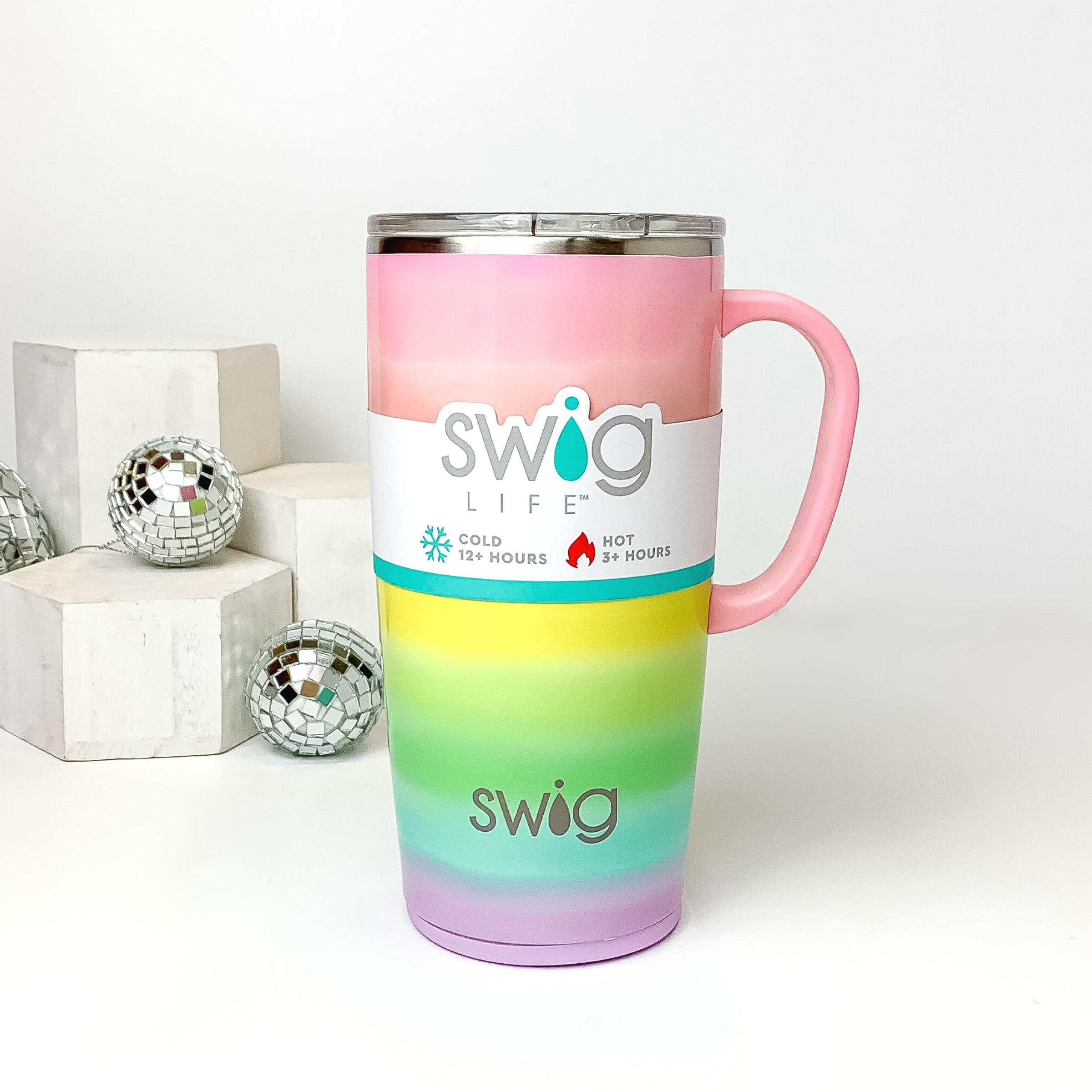 Ombre rainbow mug with a clear lid and light pink handle. This mug is pictured on a white background with white blocks and disco balls on the left side of the picture. 