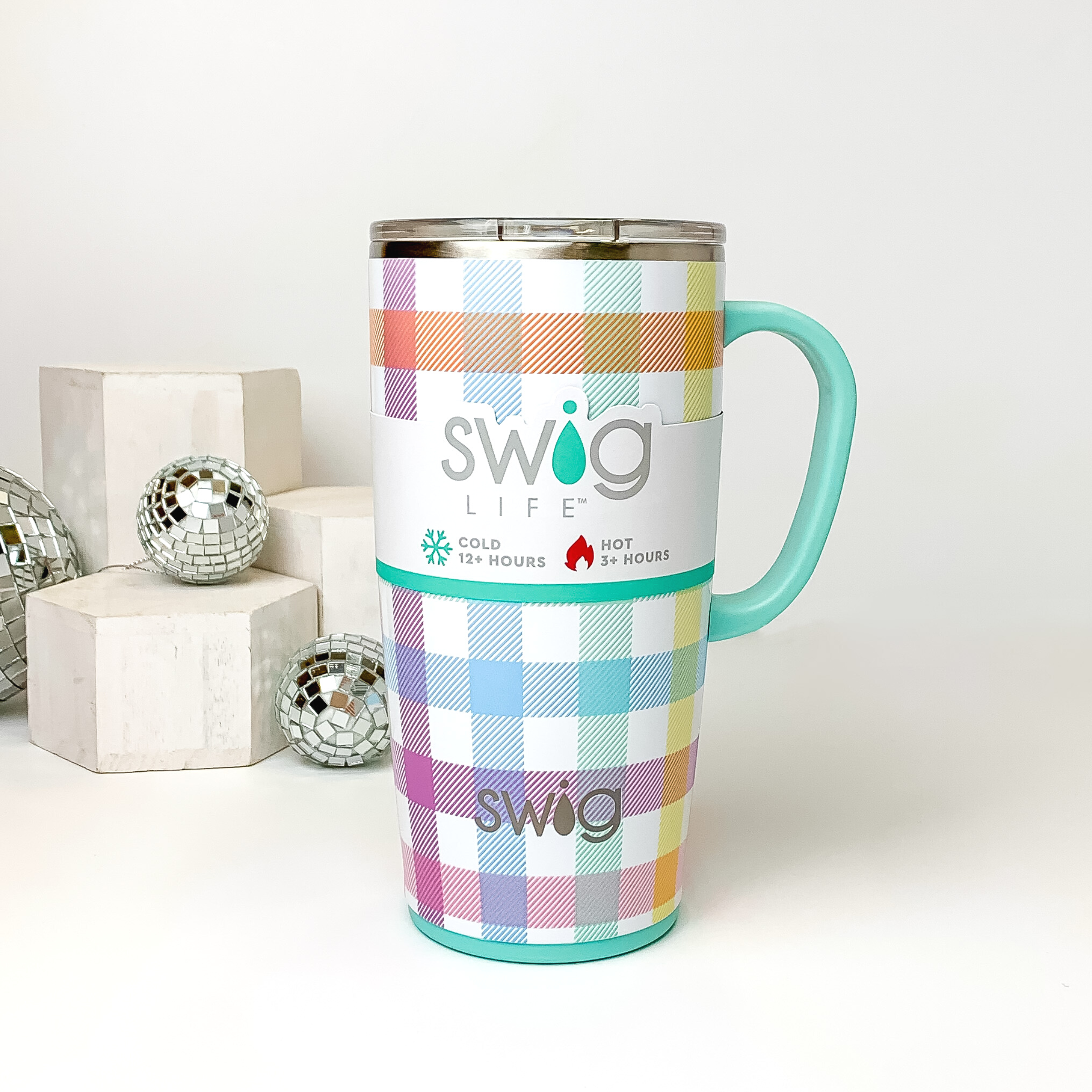 Multicolor plaid print on a white mug with a clear lid and light aqua handle. This mug is pictured on a white background with white blocks and disco balls on the left side of the picture. 