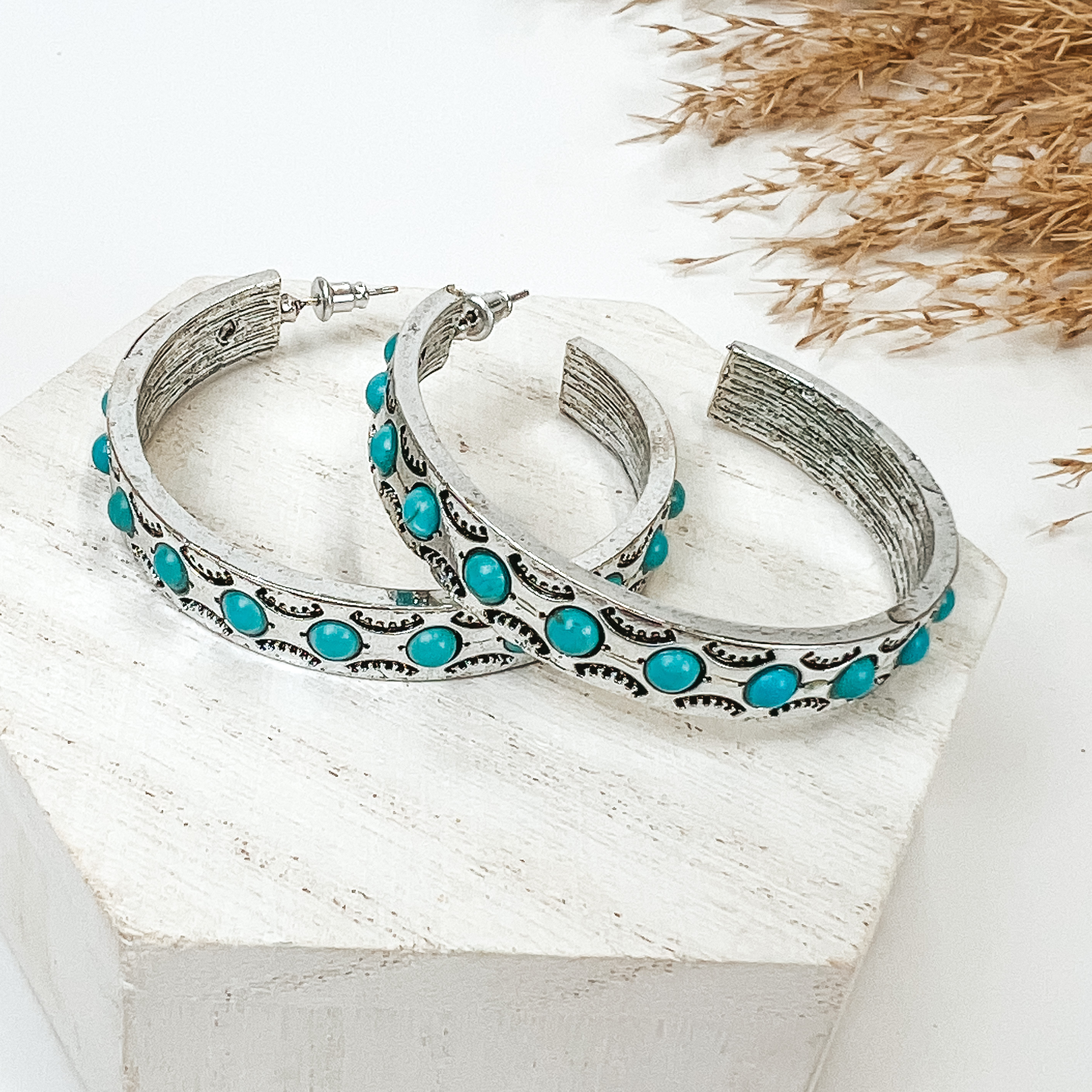 Semi hoop earrings in silver tone with turquoise stones, pictured with pampas grass, on a white background. 
