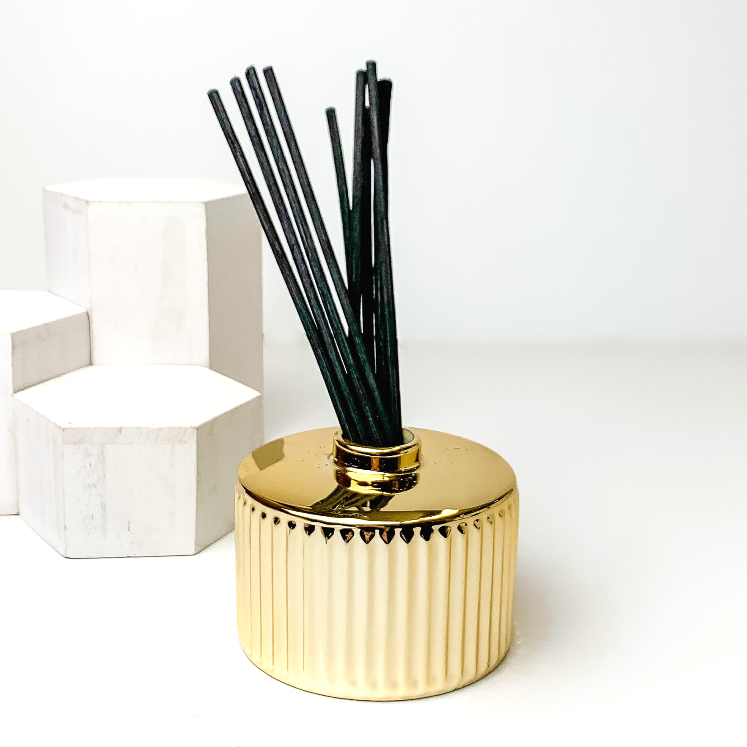 Gold diffuser holder with black reeds sticking out of the top. This diffuser is pictured on a white background with white blocks on the left side of the candle. 