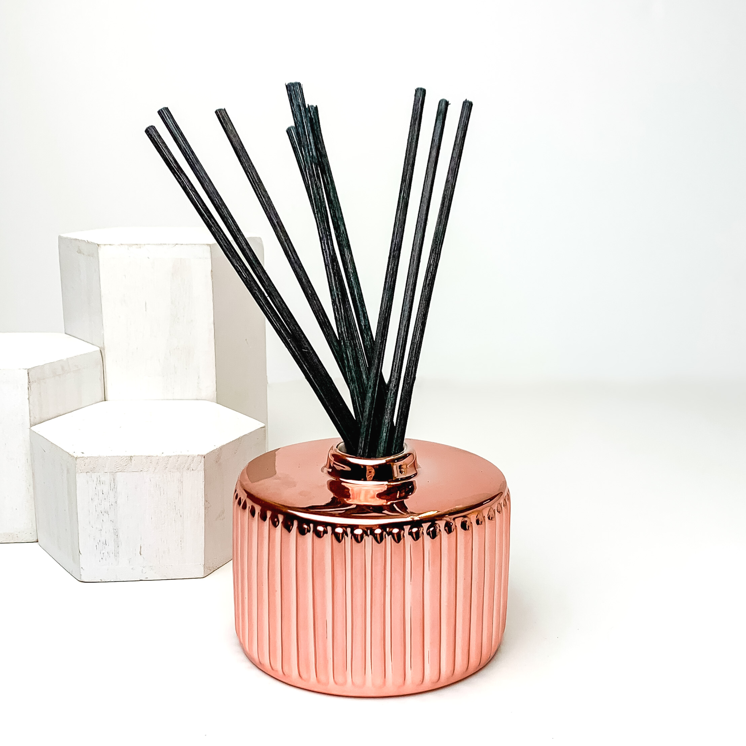 Rose gold diffuser holder with black reeds sticking out of the top. This diffuser is pictured on a white background with white blocks on the left side of the candle. 