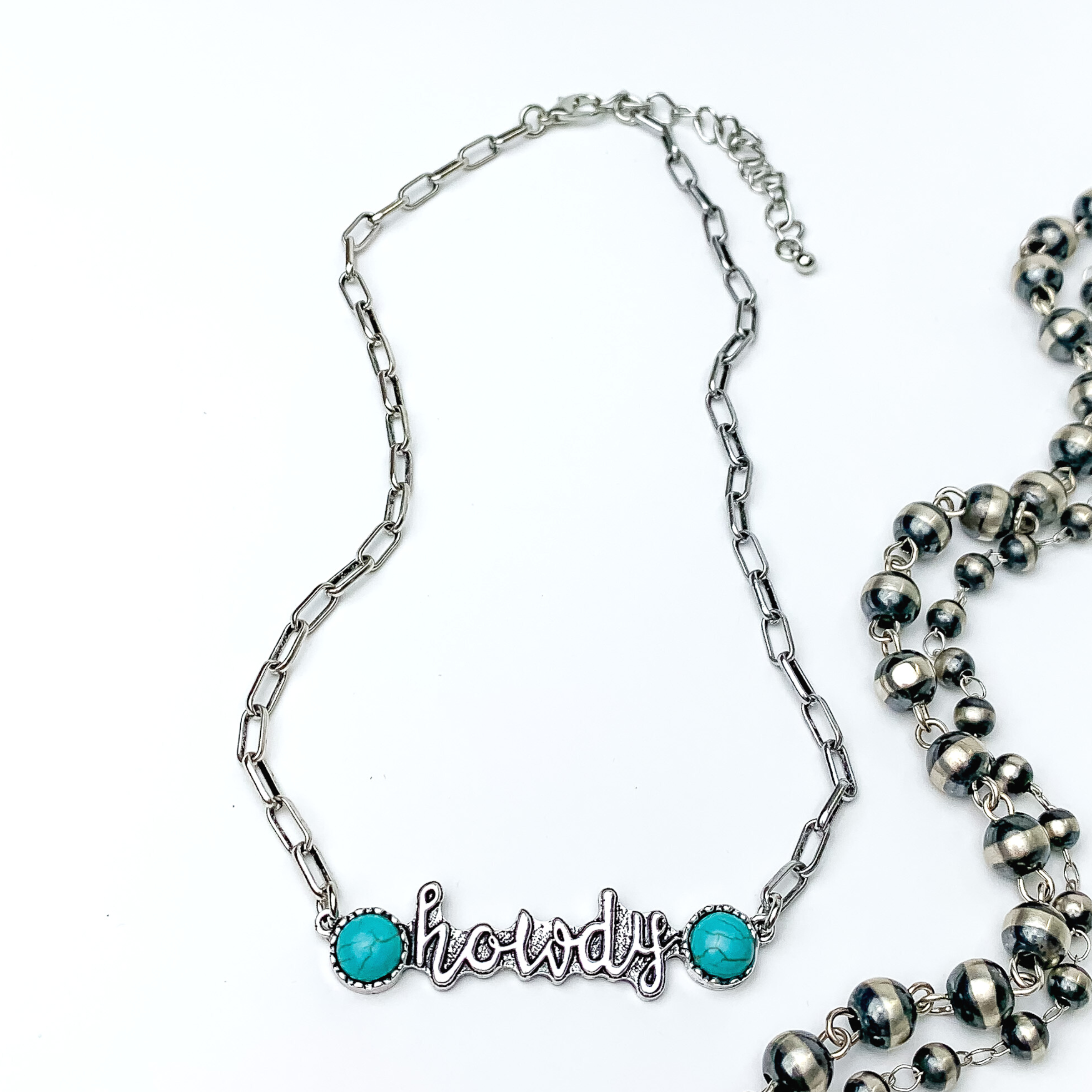 Silver chain necklace with a silver pendant that spells out "howdy" in cursive. At each end of the word is a circle, turquoise stone. This necklae is pictured on a white background with silver beads on the right side.