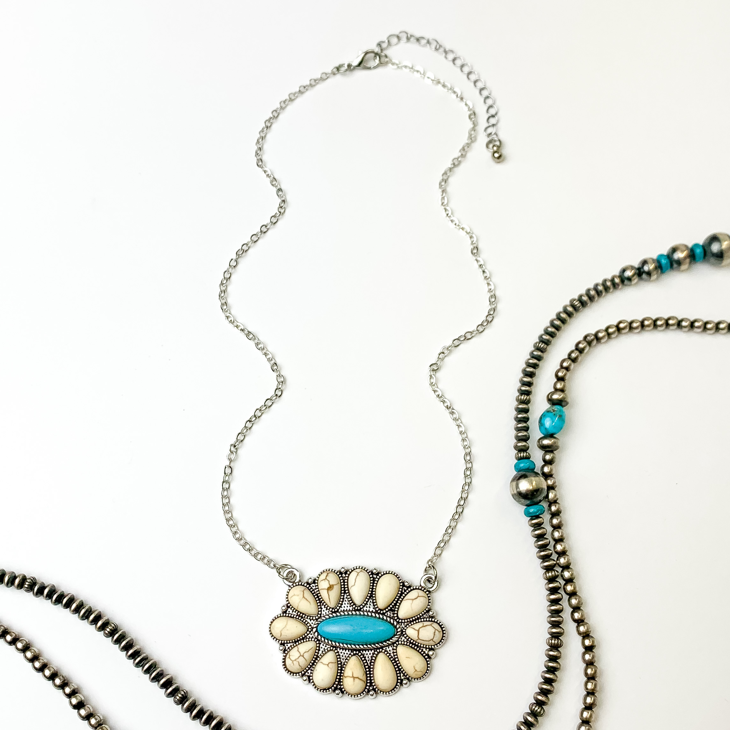 Silver chain necklace with oval cluster pendant. This pendant is mostly made up of ivory stones with a center turquoise stone. This necklace is pictured on an white background with silver and turquoise beads on the right side of the picture.