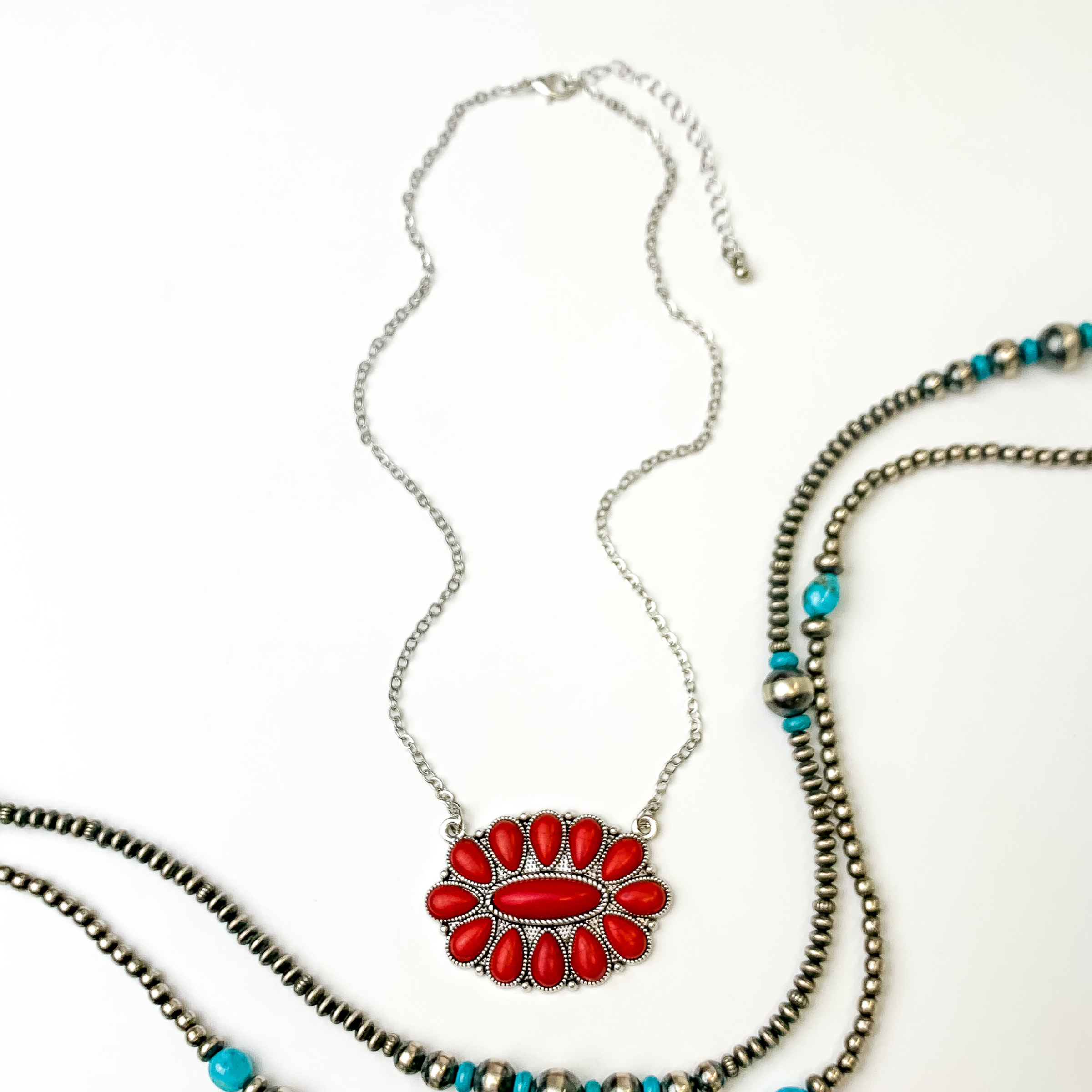 Silver chain necklace with oval cluster pendant. This pendant is made up of red stones. This necklace is pictured on an white background with silver and turquoise beads on the right side of the picture.