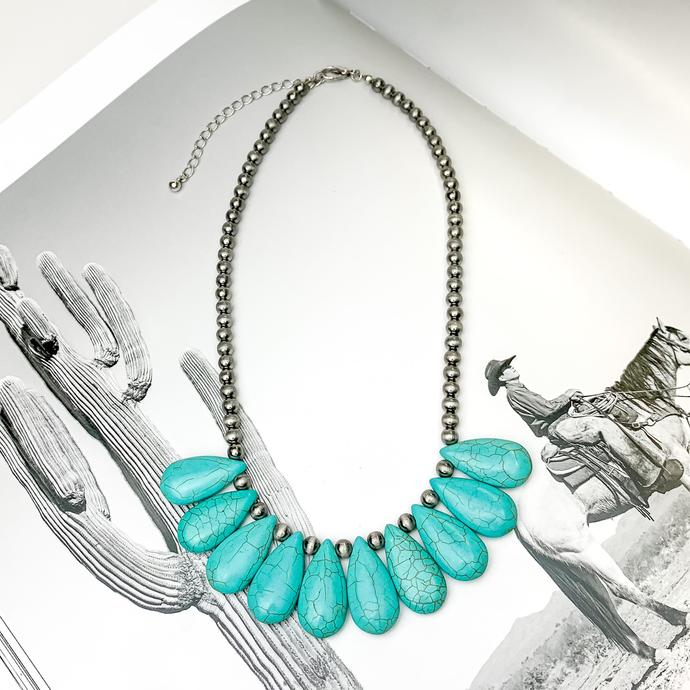 Silver beaded necklace with ten turquoise teardrop beads. This necklace is pictured on an open black and white western picture book. 