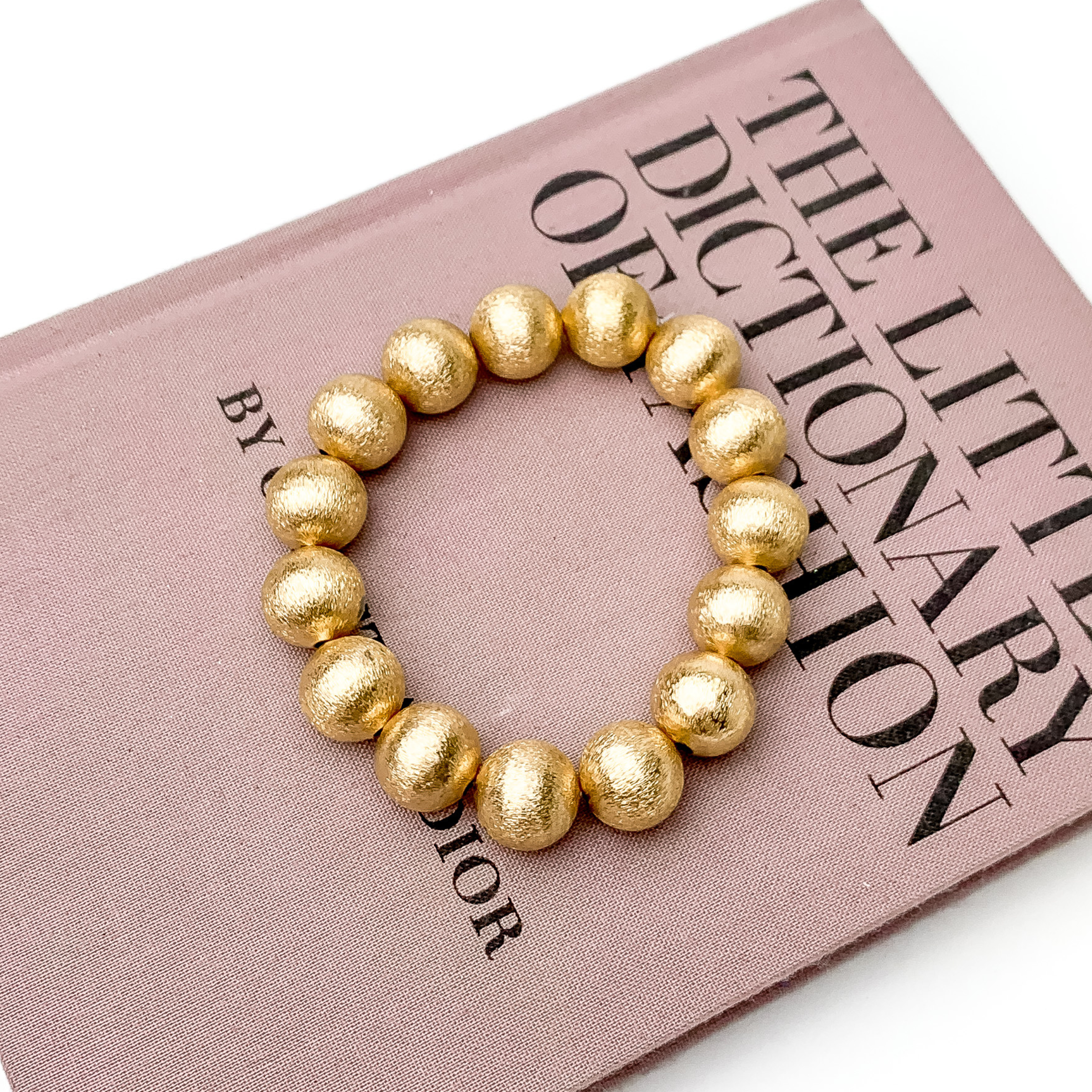 Gold beaded bracelet pictured laying on a mauve book on a white background.  