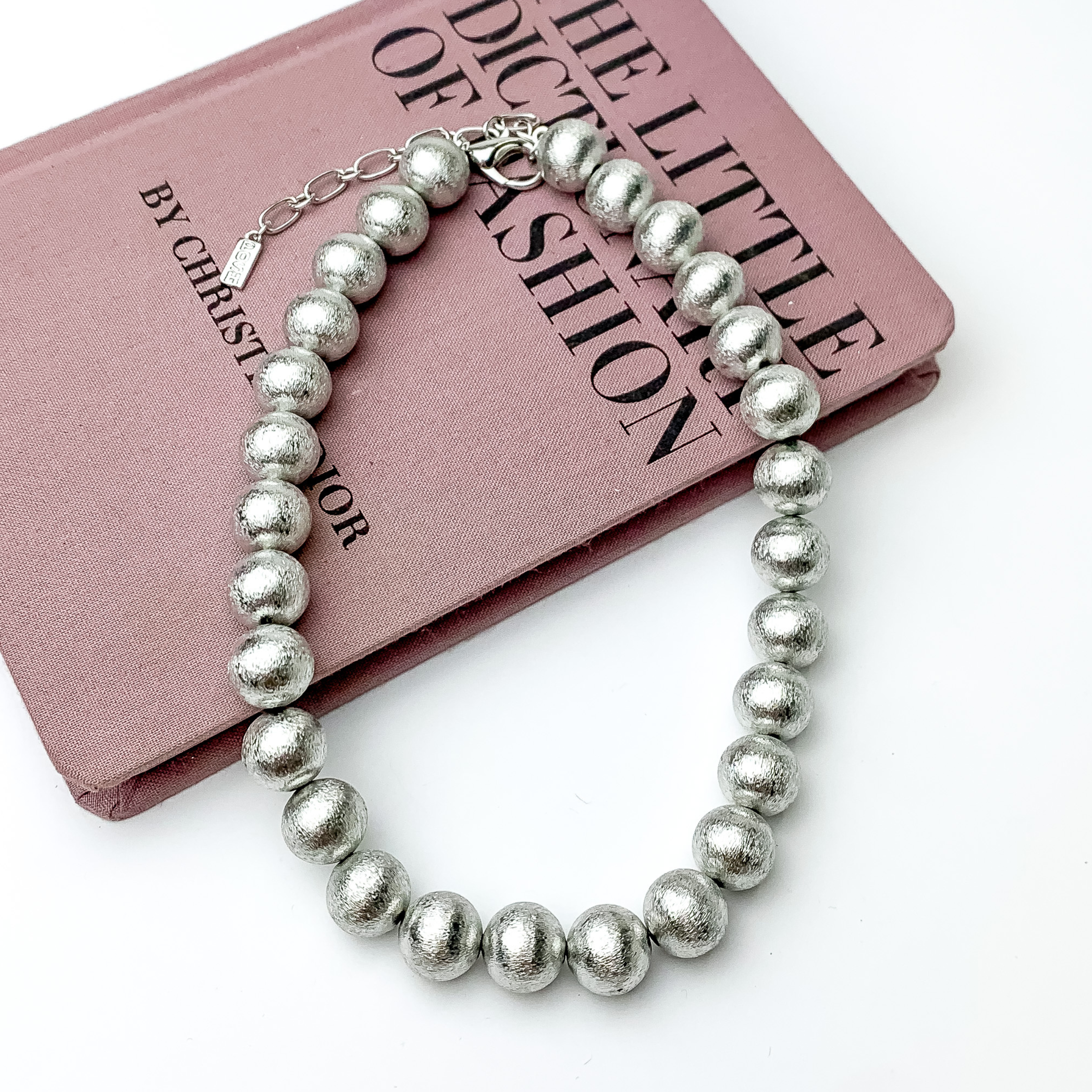 Silver beaded necklace pictured laying partially on a mauve book on a white background.
