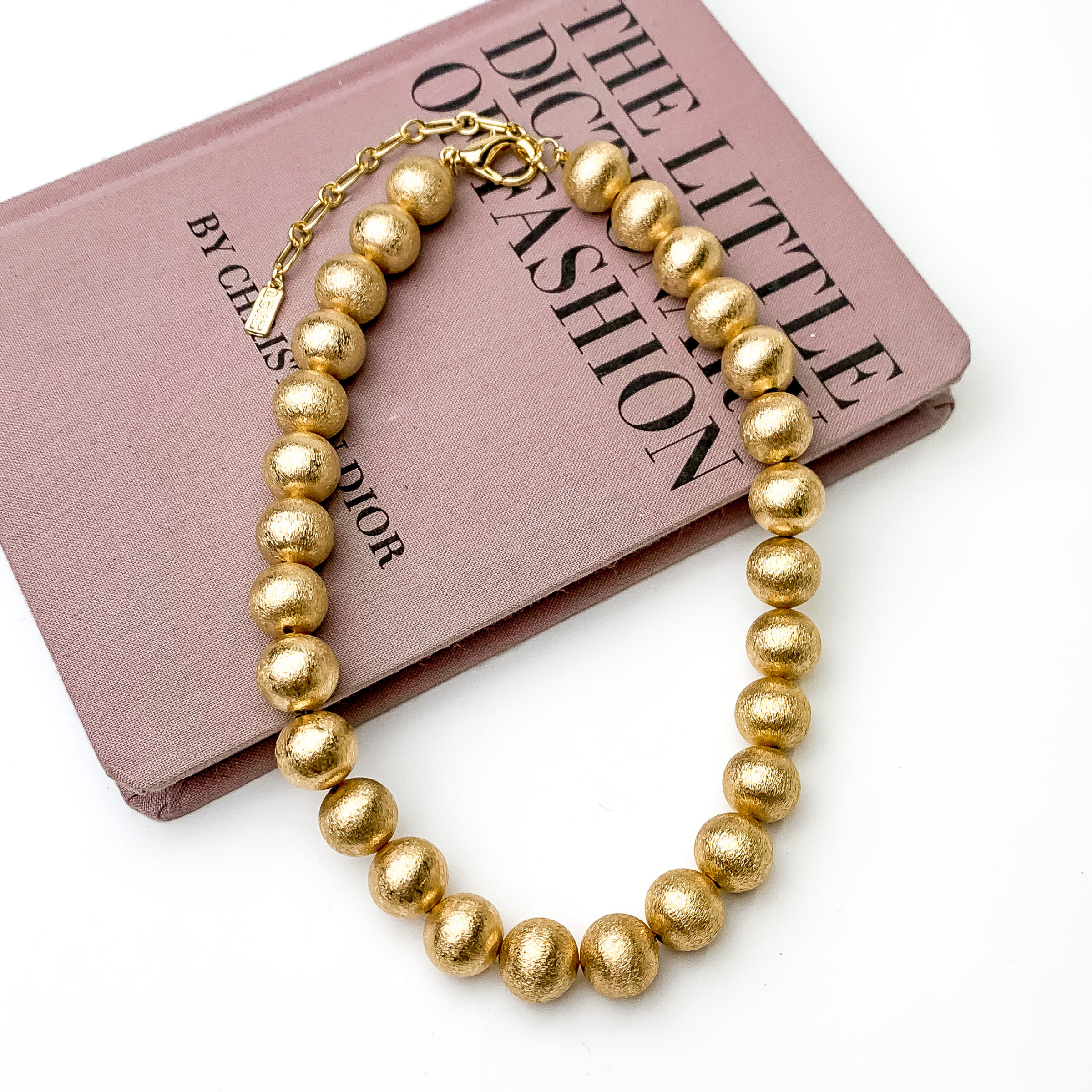 Gold beaded necklace pictured laying partially on a mauve book on a white background. 