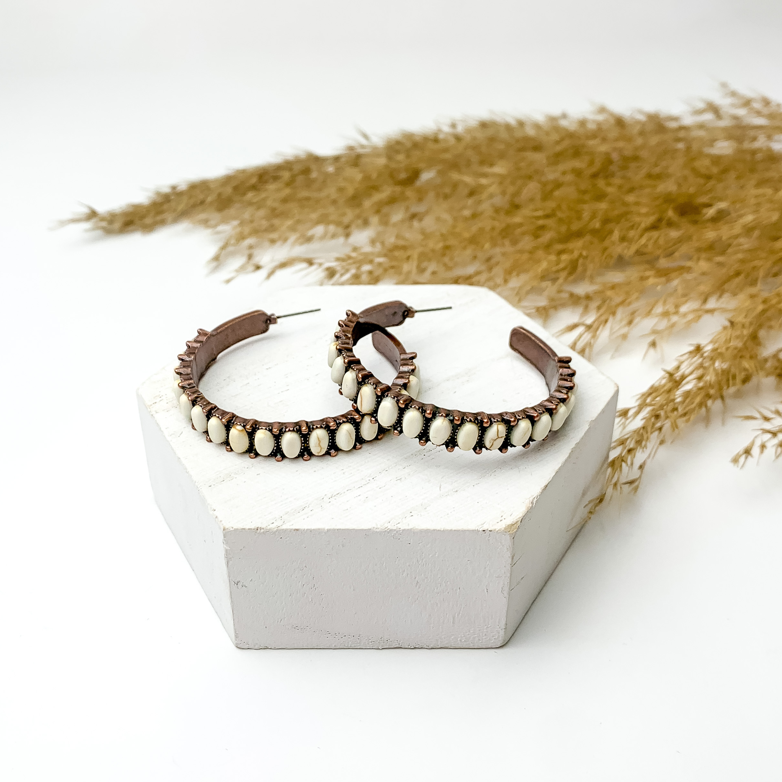 Copper tone semi hoop earrings with white stones, pictured with pampas grass on a white background. 