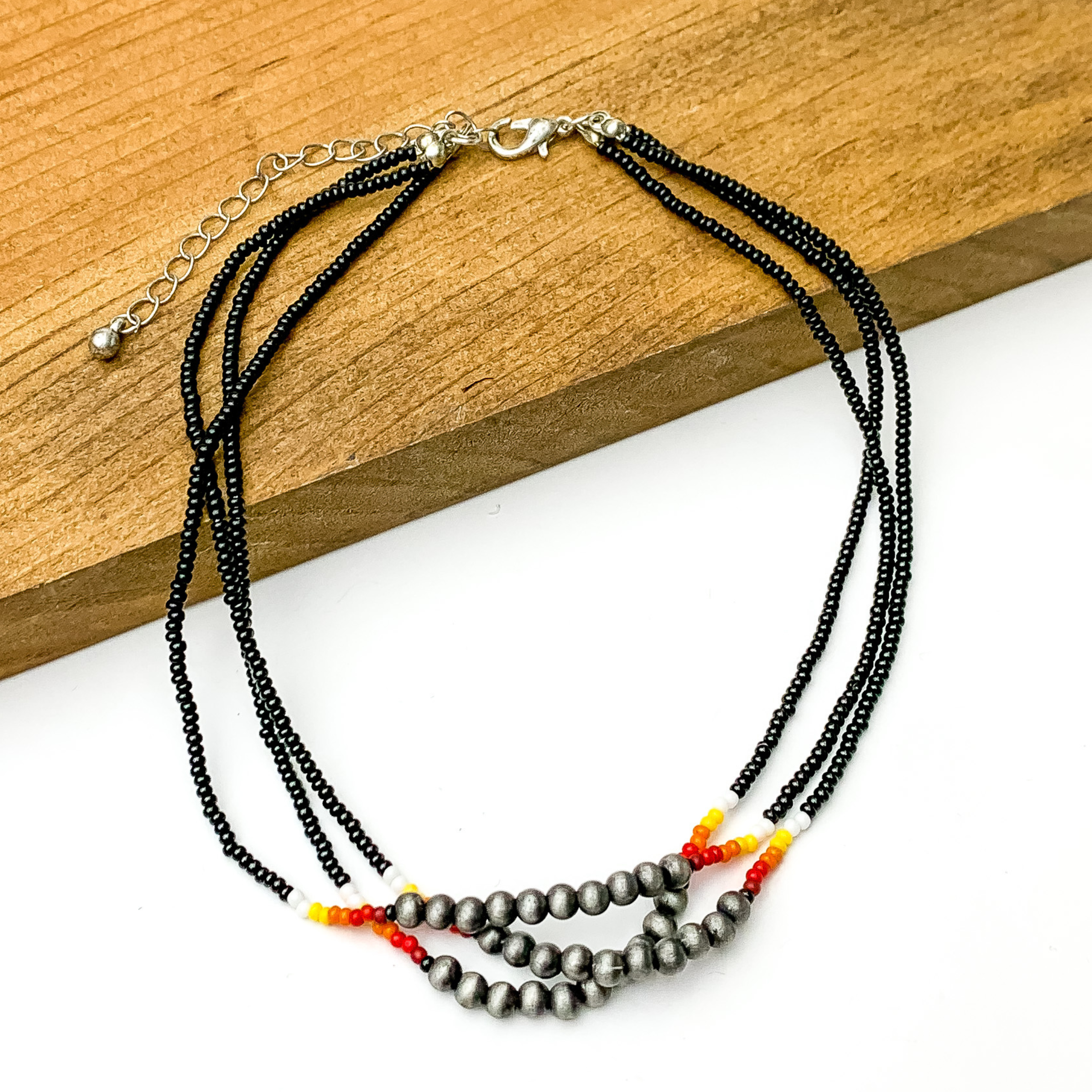 Three stranded necklace with mostly black beads. This necklace also includes a segment of silver beads on each strand with a couple white, red, and orange beads on each side of the segment. This necklace is pictured partially on a block of wood on a white background.  