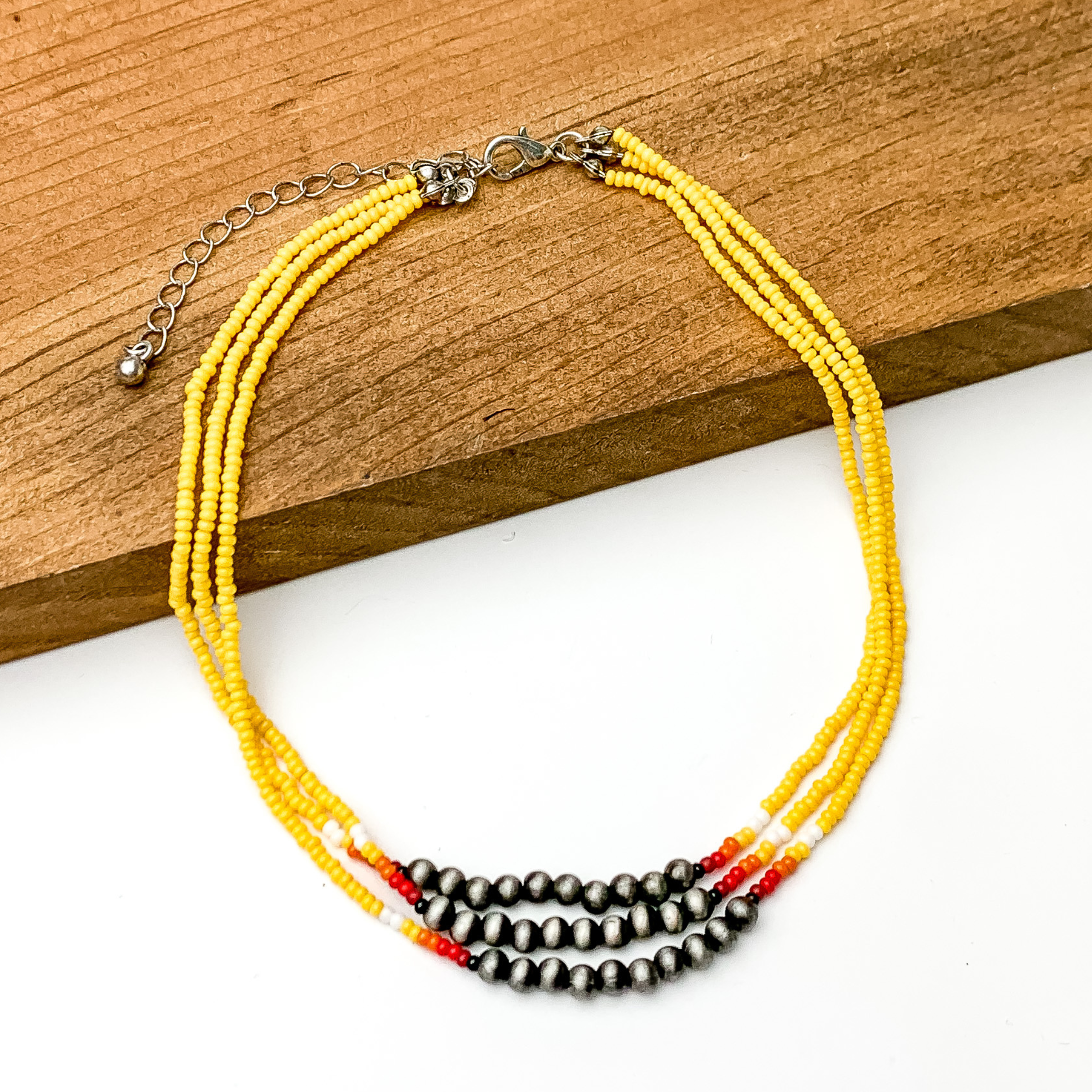 Three stranded necklace with mostly yellow beads. This necklace also includes a segment of silver beads on each strand with a couple white, red, and orange beads on each side of the segment. This necklace is pictured partially on a block of wood on a white background.  