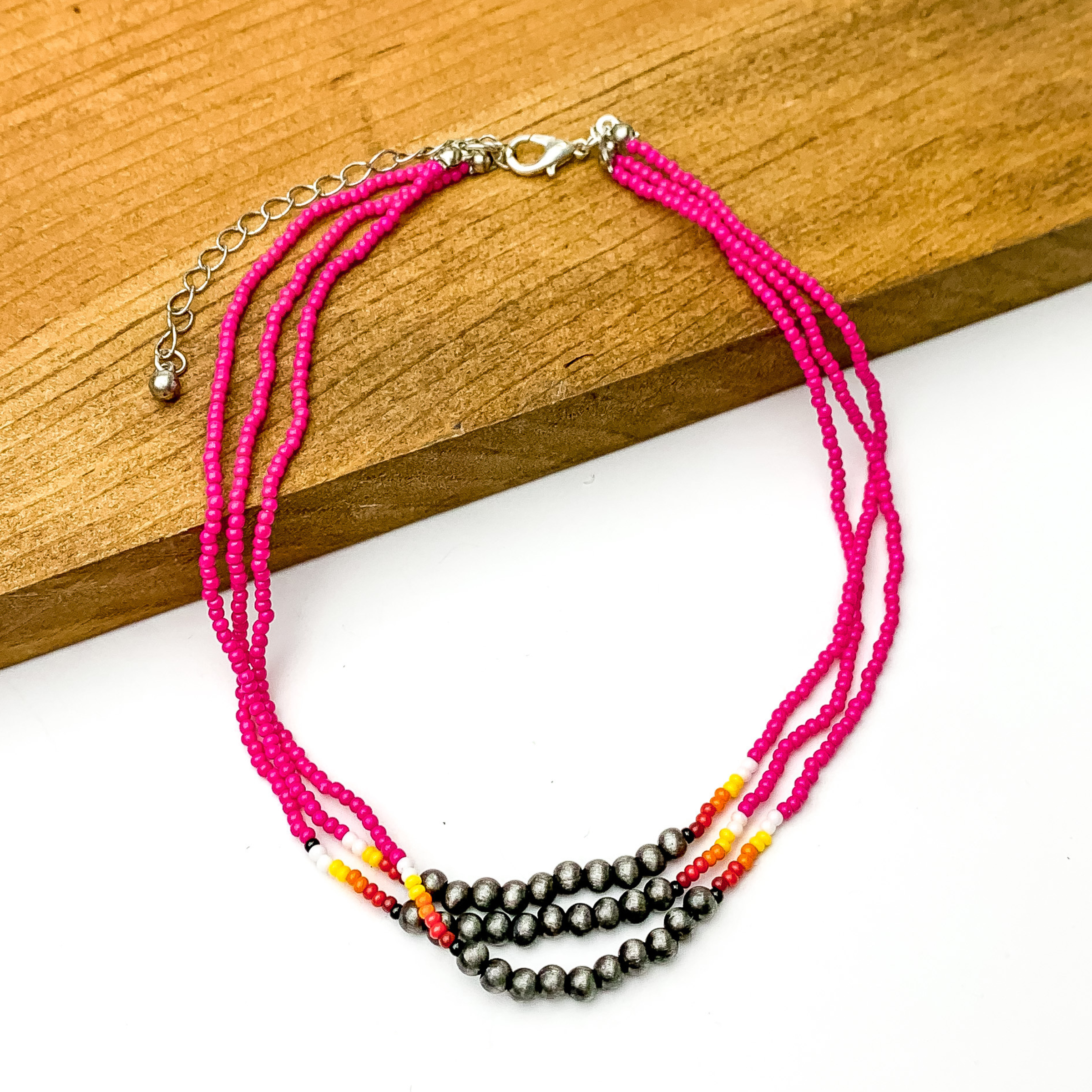 Three stranded necklace with mostly pink beads. This necklace also includes a segment of silver beads on each strand with a couple white, red, and orange beads on each side of the segment. This necklace is pictured partially on a block of wood on a white background.  