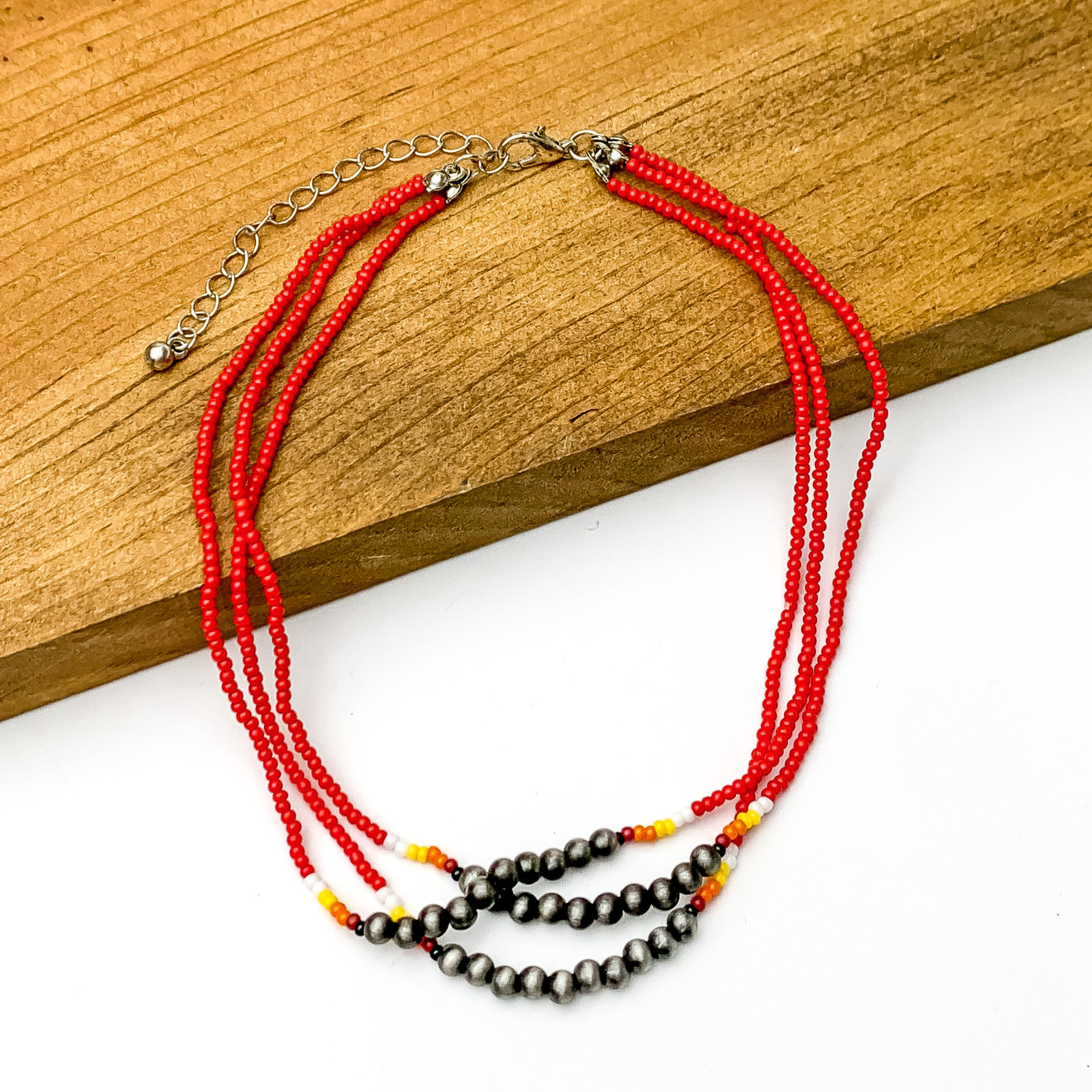 Three stranded necklace with mostly red beads. This necklace also includes a segment of silver beads on each strand with a couple white, red, and orange beads on each side of the segment. This necklace is pictured partially on a block of wood on a white background.  