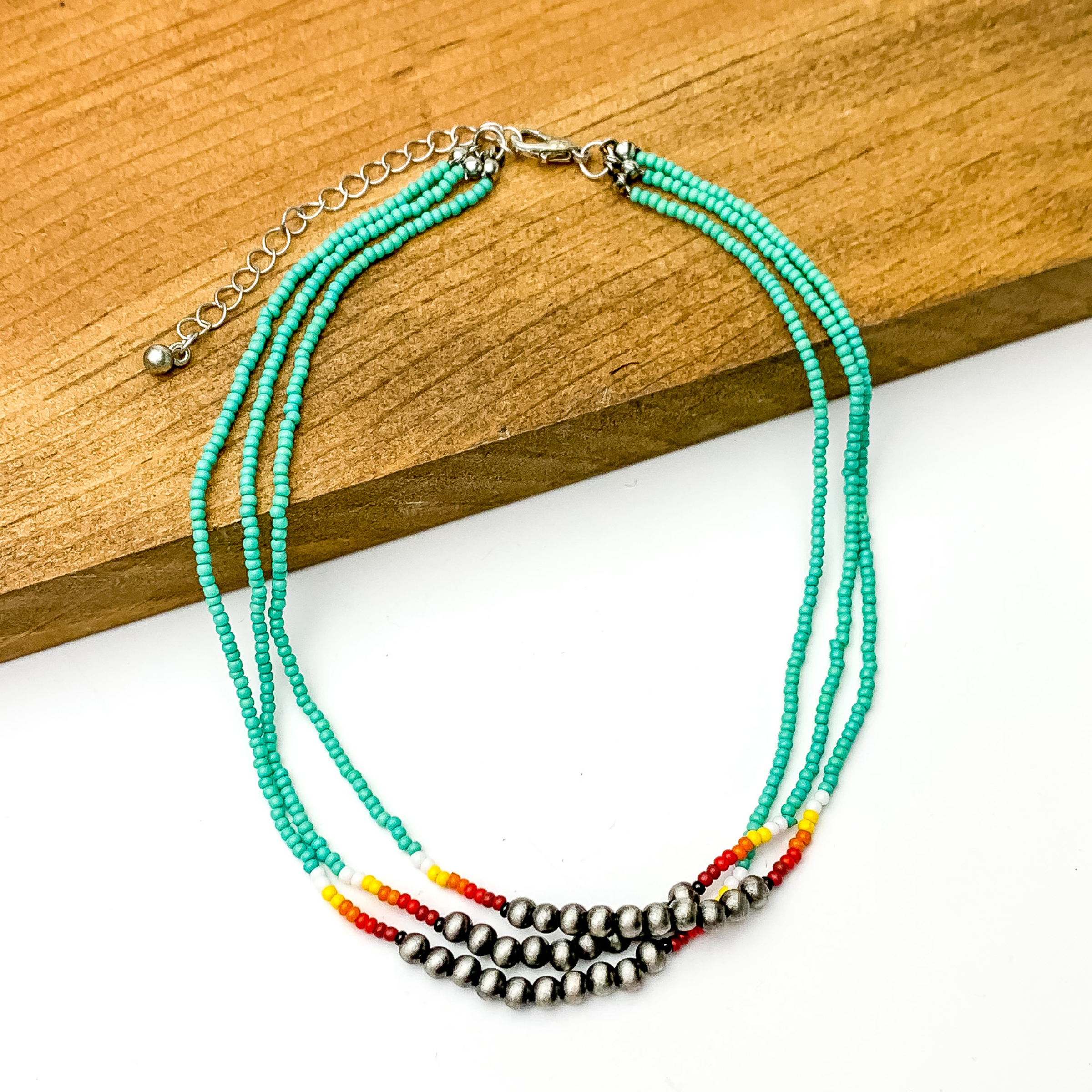 Three stranded necklace with mostly turquoise beads. This necklace also includes a segment of silver beads on each strand with a couple white, red, and orange beads on each side of the segment. This necklace is pictured partially on a block of wood on a white background.  