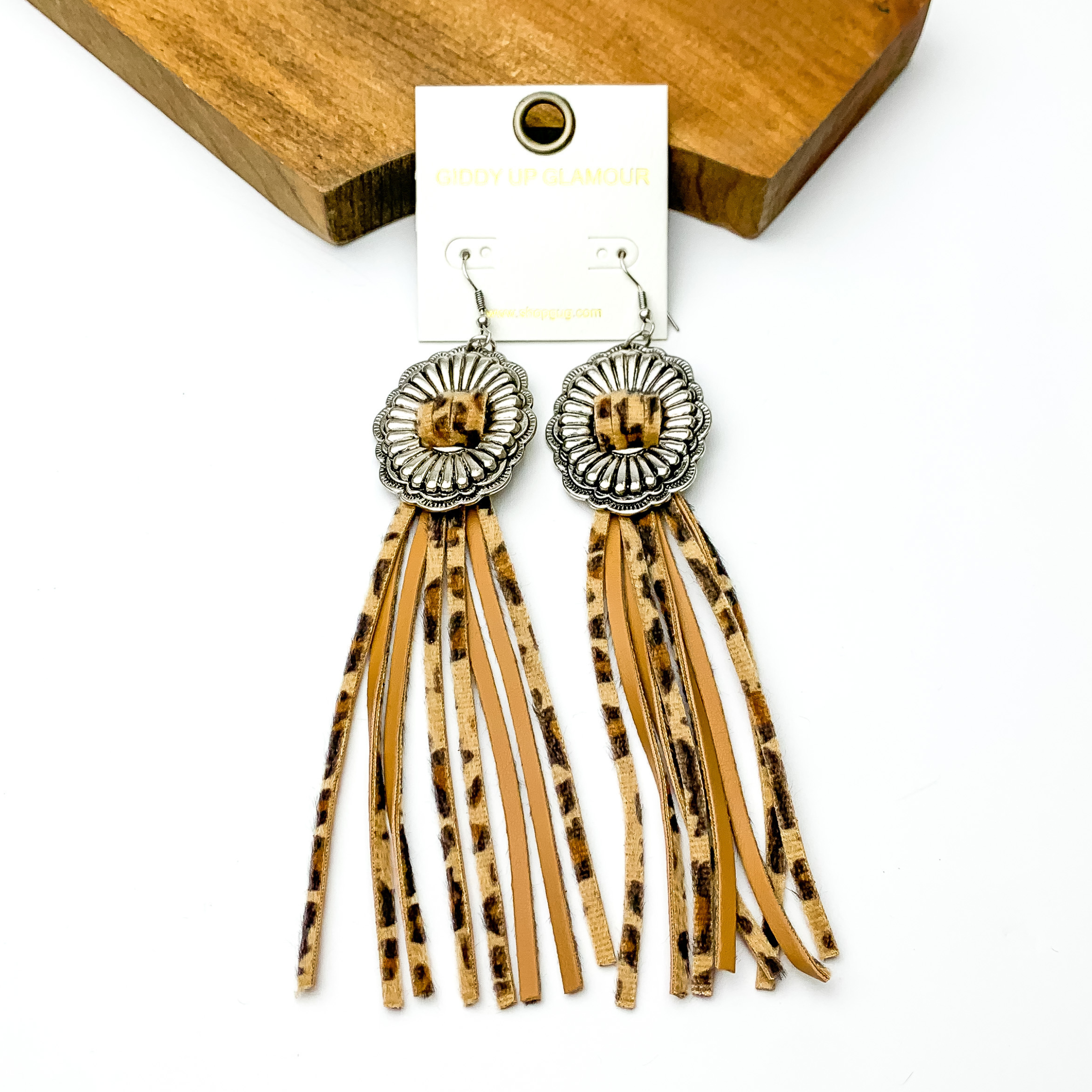 Silver concho dangle earrings with leopard print tassels. These earrings are pictured on a white background with a brown block at the top of the picture. 