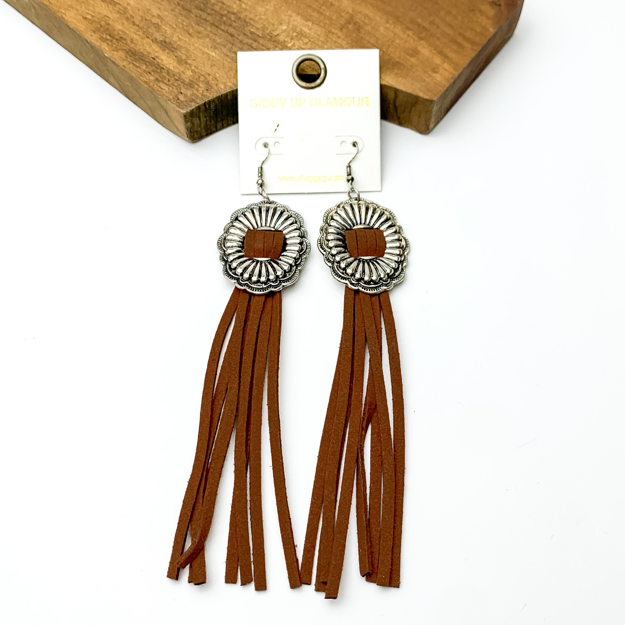 Silver concho dangle earrings with brown tassels. These earrings are pictured on a white background with a brown block at the top of the picture. 