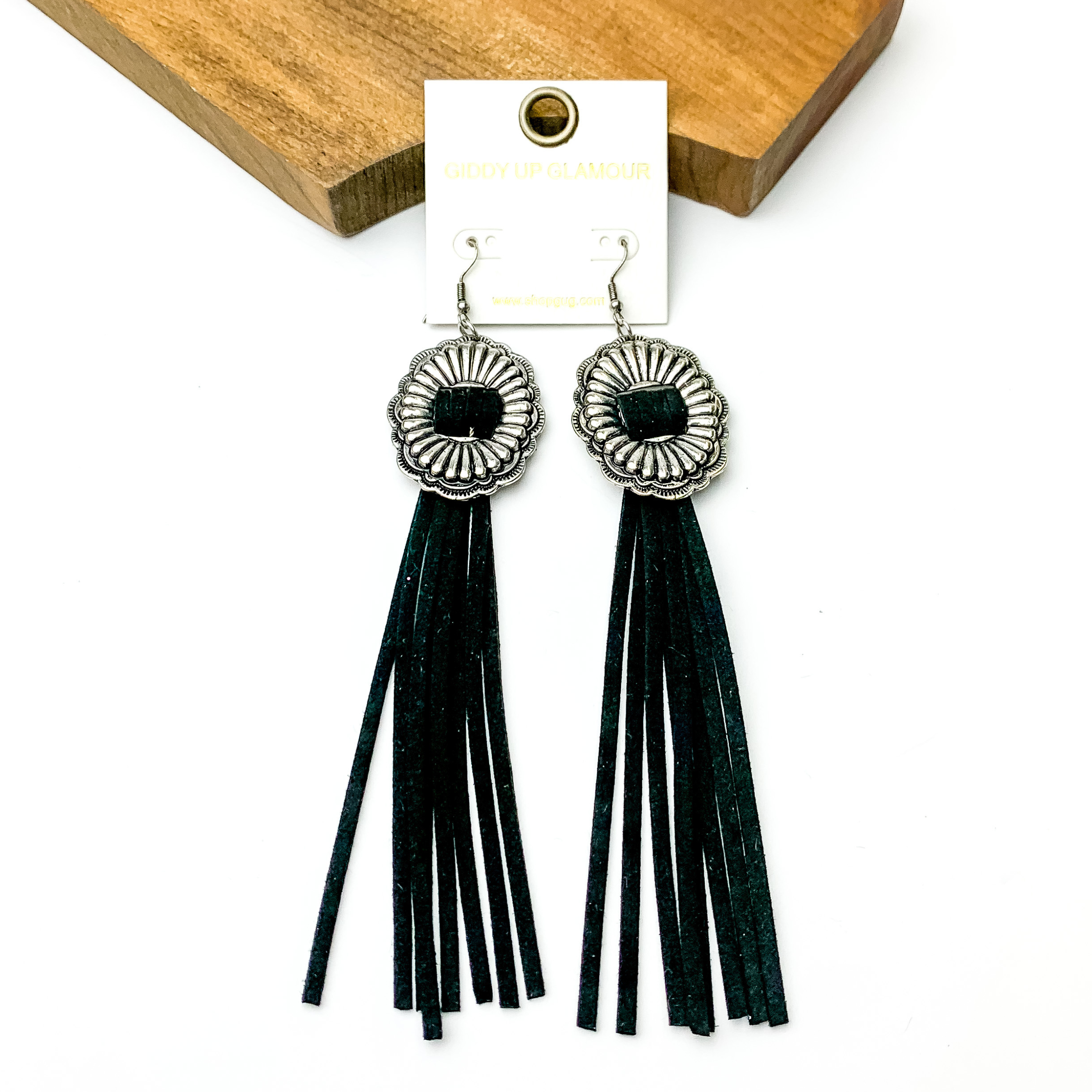 Silver concho dangle earrings with black tassels. These earrings are pictured on a white background with a brown block at the top of the picture. 