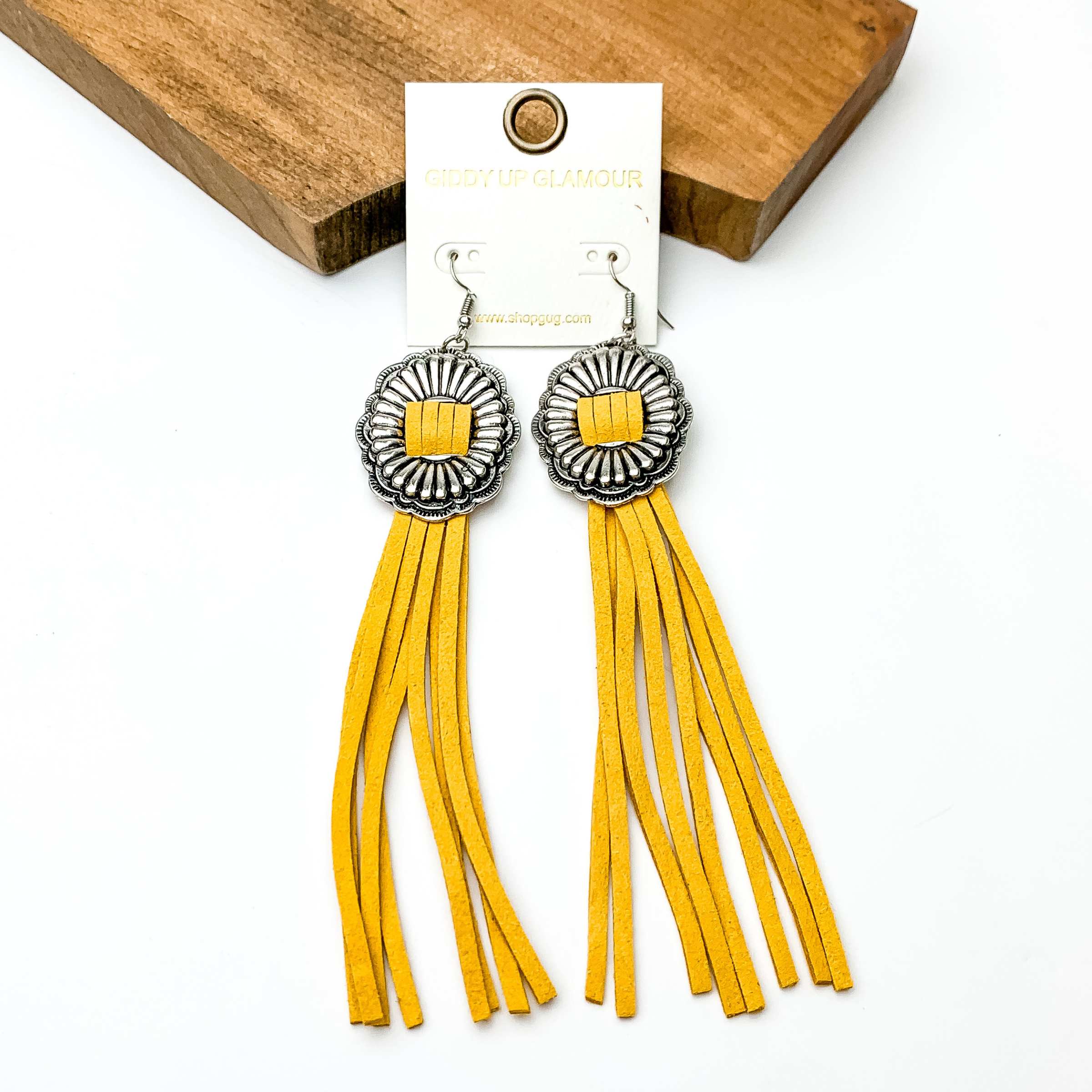 Silver concho dangle earrings with yellow tassels. These earrings are pictured on a white background with a brown block at the top of the picture. 