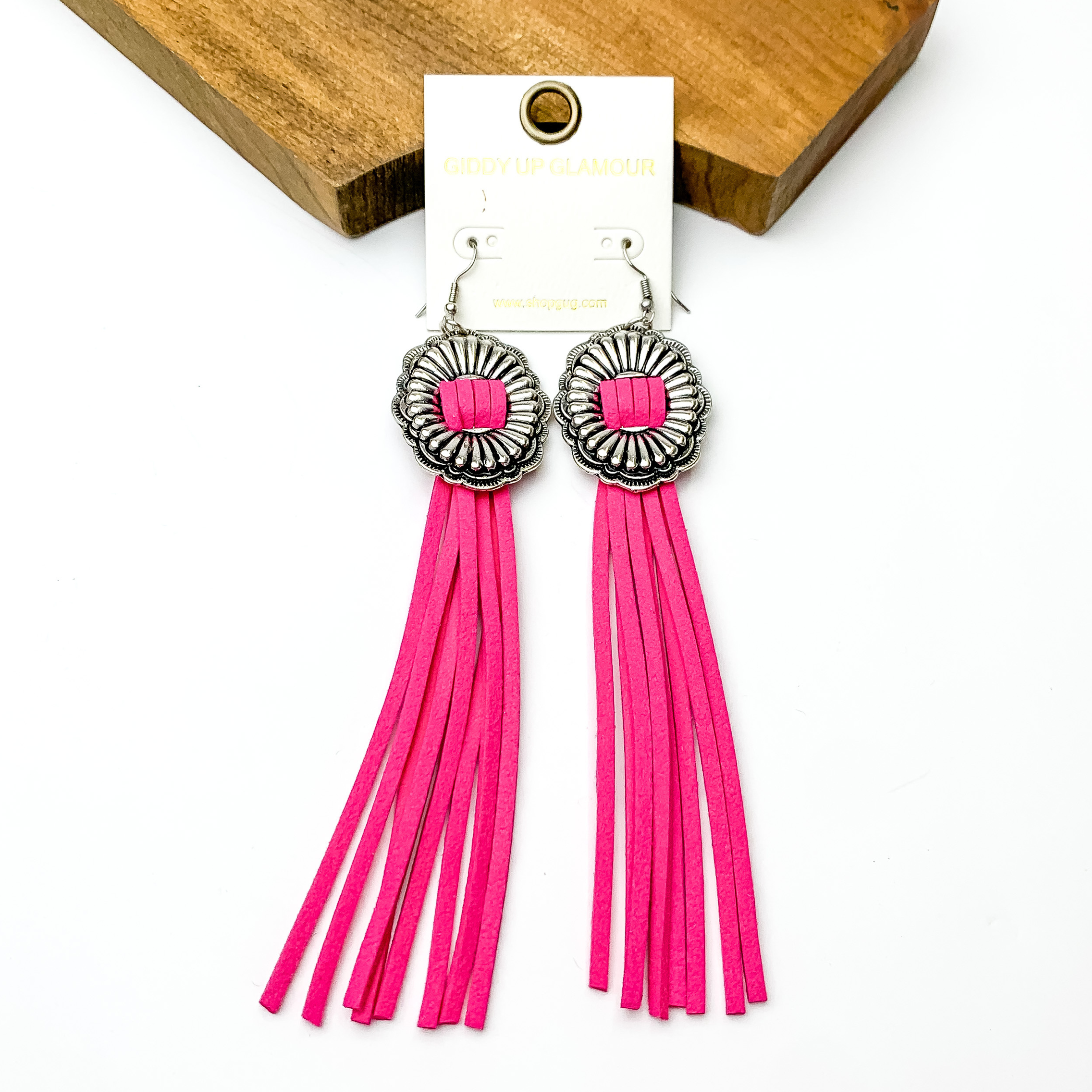 Silver concho dangle earrings with pink tassels. These earrings are pictured on a white background with a brown block at the top of the picture. 