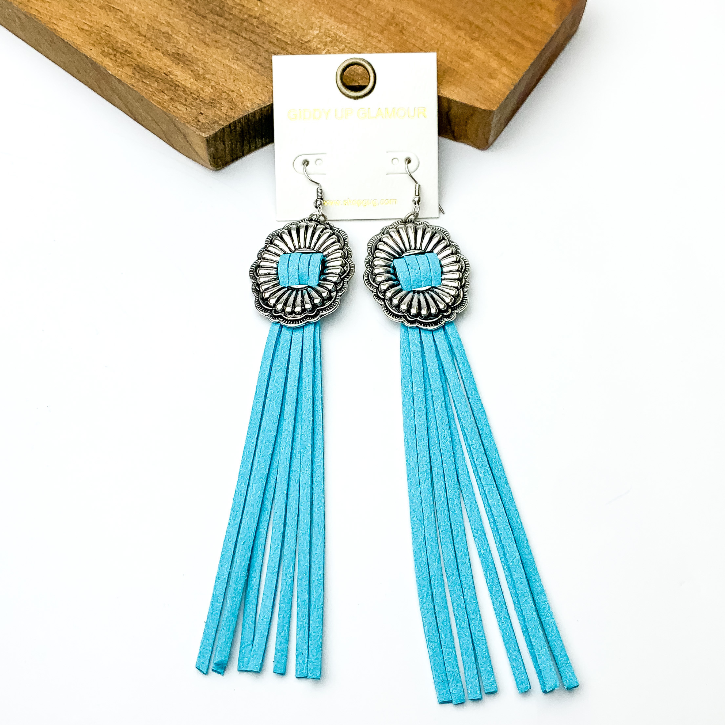 Silver concho dangle earrings with light blue tassels. These earrings are pictured on a white background with a brown block at the top of the picture. 