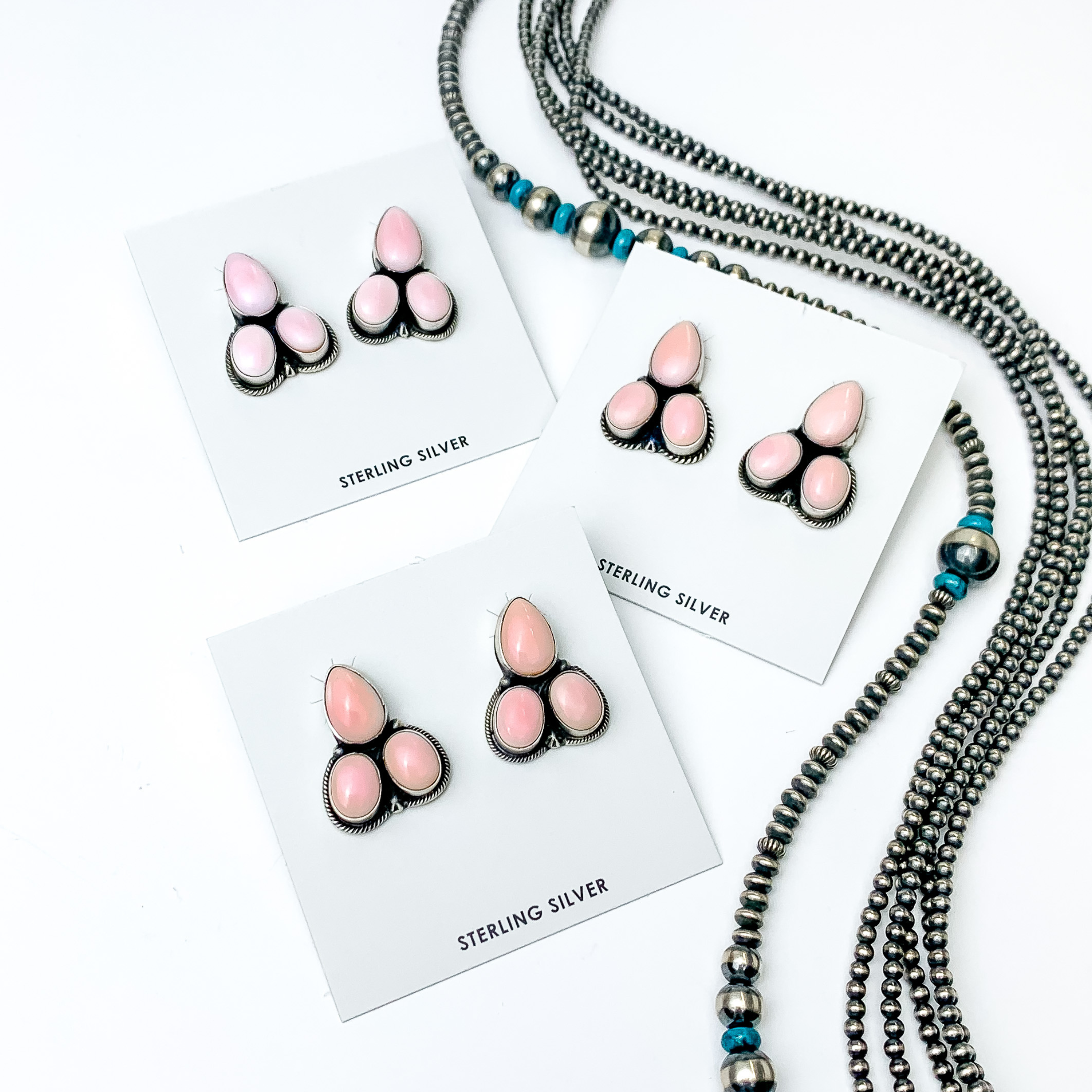 Pictured are three pairs of pink, stone stud earrings with silver outline. The earrings include a teardrop stone and two circle stones beneath the teardrop stone. These earrings are pictured on a white background with silver beads on the right side of the picture. 
