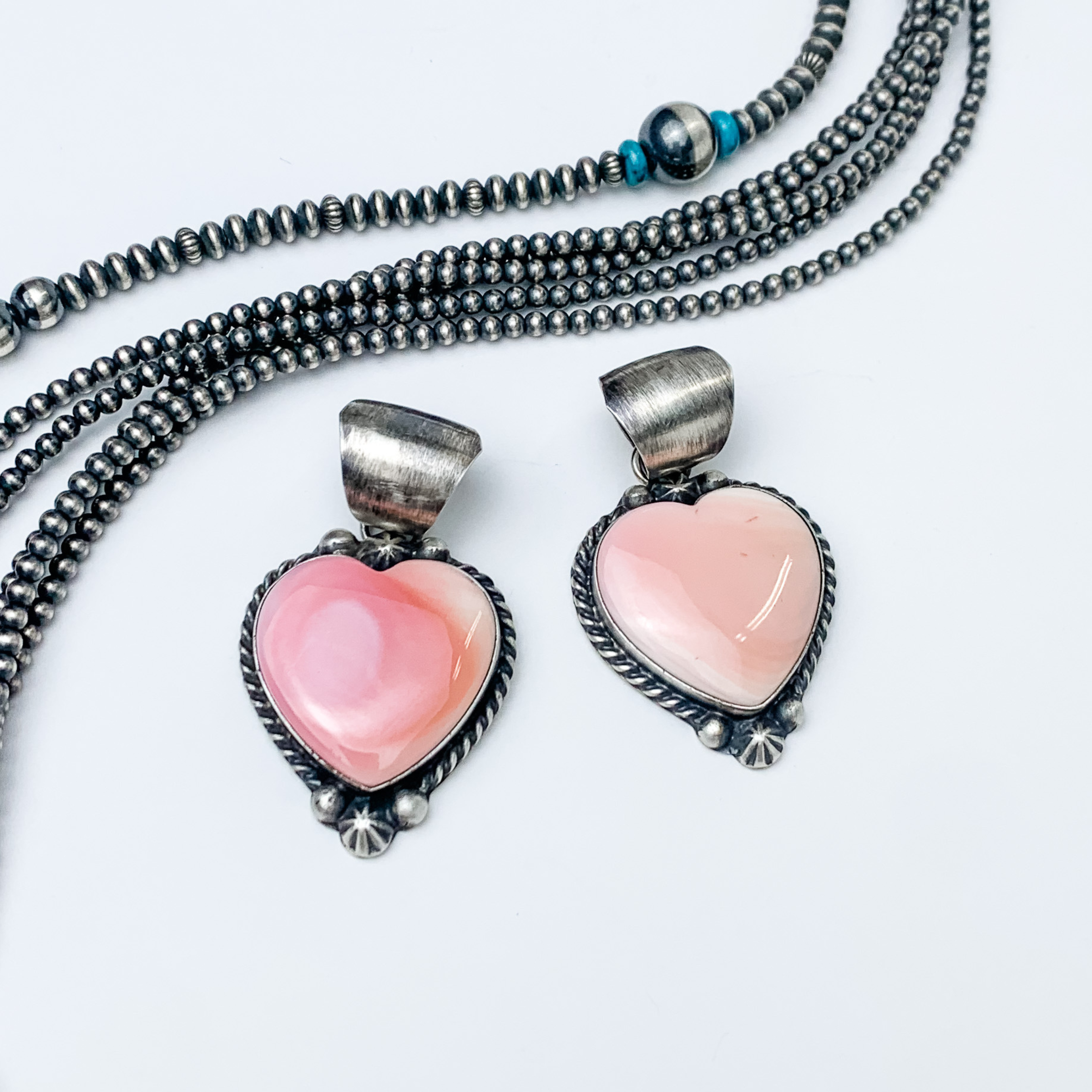 Pictured are two pink heart stone pendants with a silver outline and silver bail.  These pendants are pictured on a white background with silver beads above the pendant. 