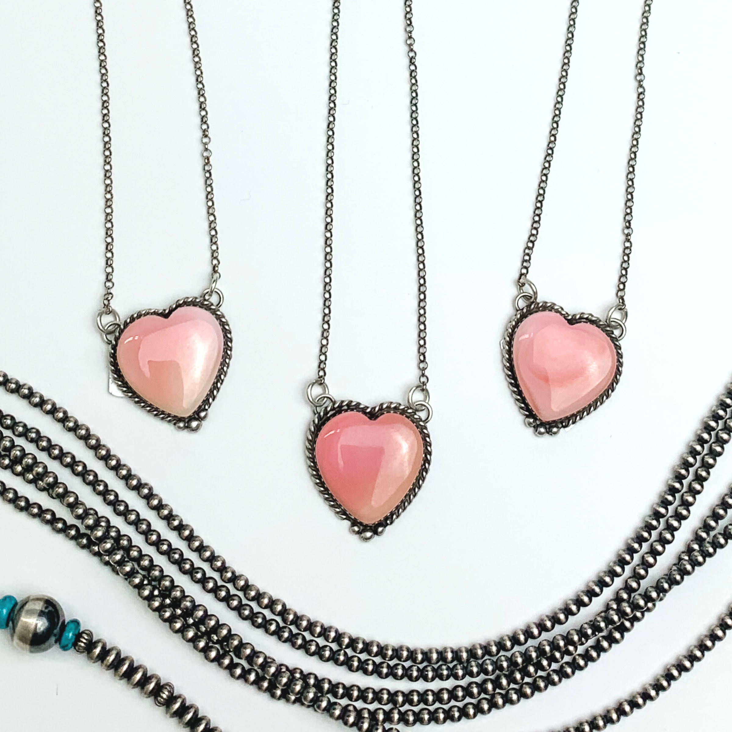 Pictured are three pink conch heart pendant necklaces. These necklaces include a silver outline and silver chain necklace. These necklaces are pictured on a white background with silver beads at the bottom of the picture. 