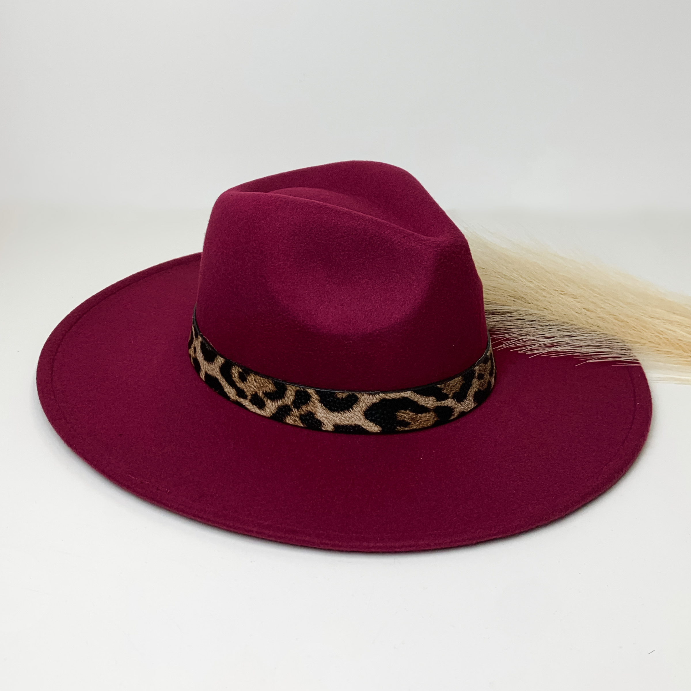 Burgundy flat brim hat with a leopard print hat bad around the crown. This hat is pictured on a white background with ivory pompous grass laying partially on the brim. 
