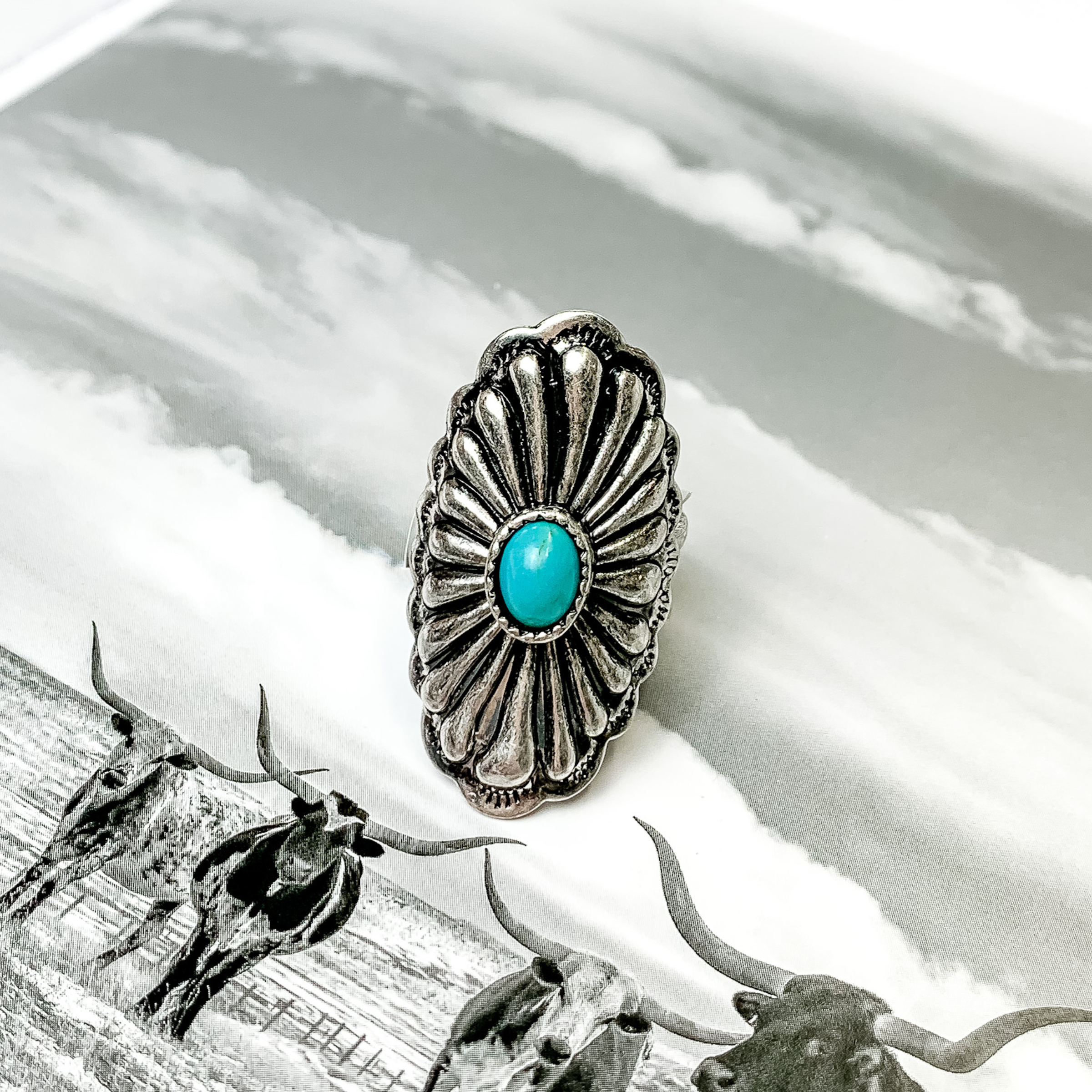 Silver, oval concho ring with a small turquoise center stone. This ring is pictured on a black and white picture. 