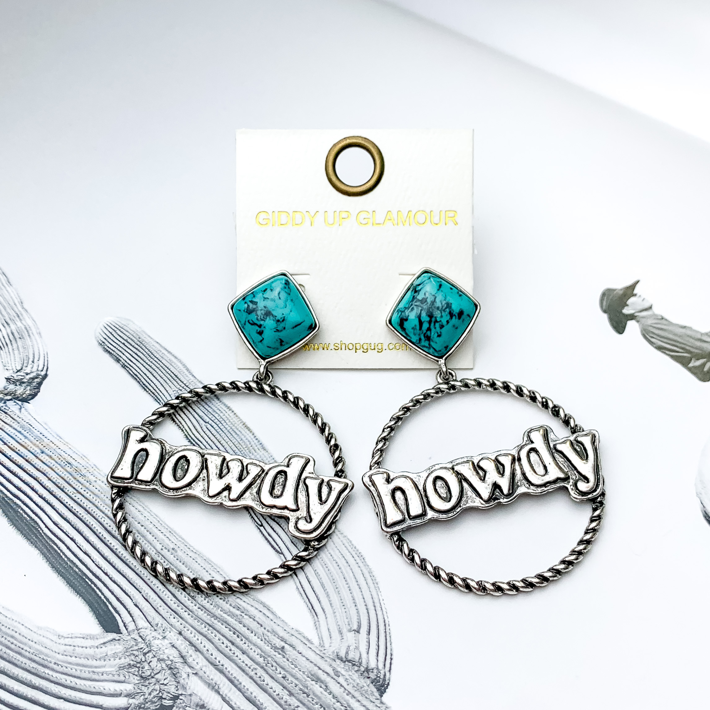 Square turquoise stud earrings with a silver rope hoop drop. In the center of the hoop, there is an engraved pendant that has the word "howdy" on it. These earrings are pictured on a black and white picture. 