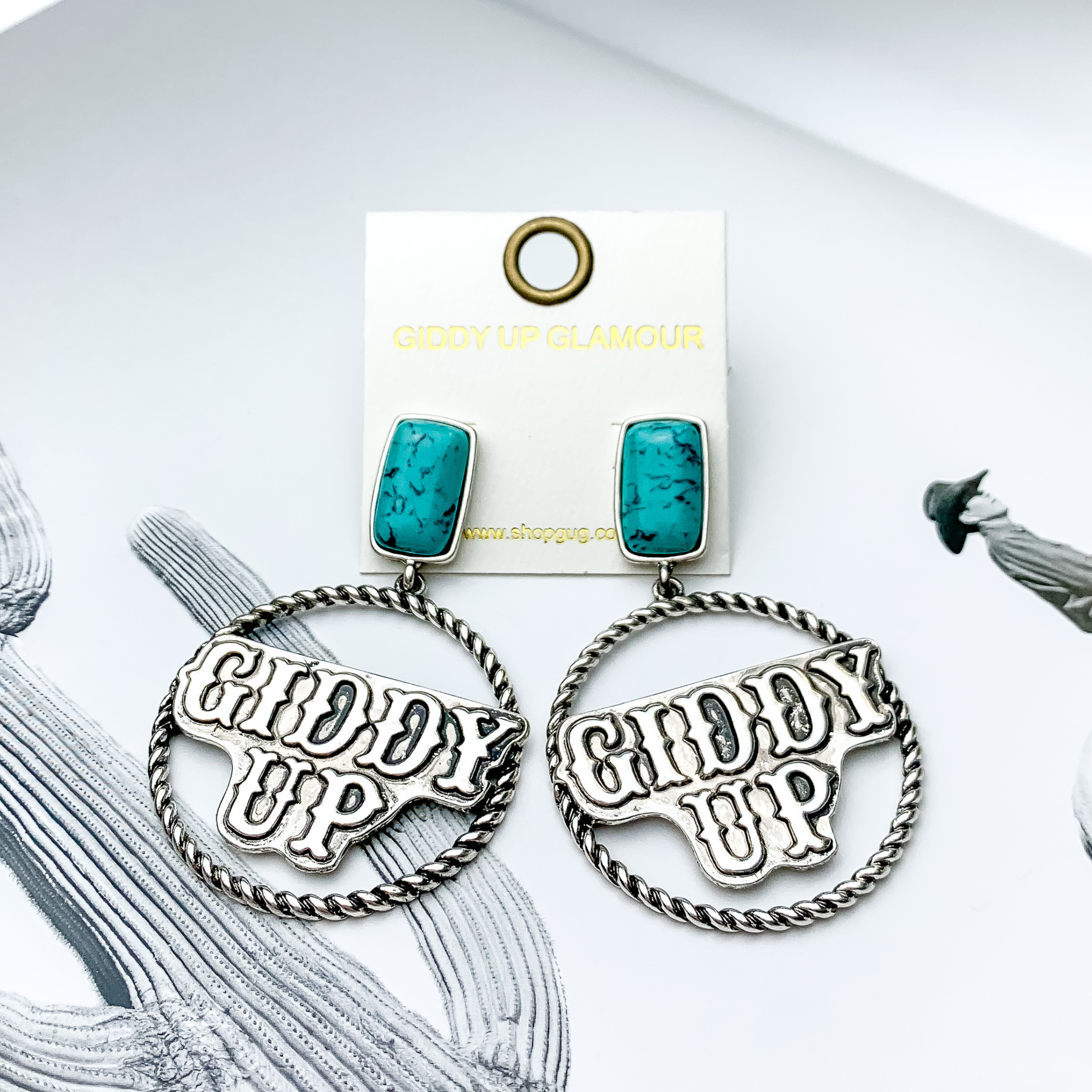 Rectangle turquoise stud earrings with a silver rope hoop drop. In the center of the hoop, there is an engraved pendant that has the words "GIDDY UP" on it. These earrings are pictured on a black and white picture. 