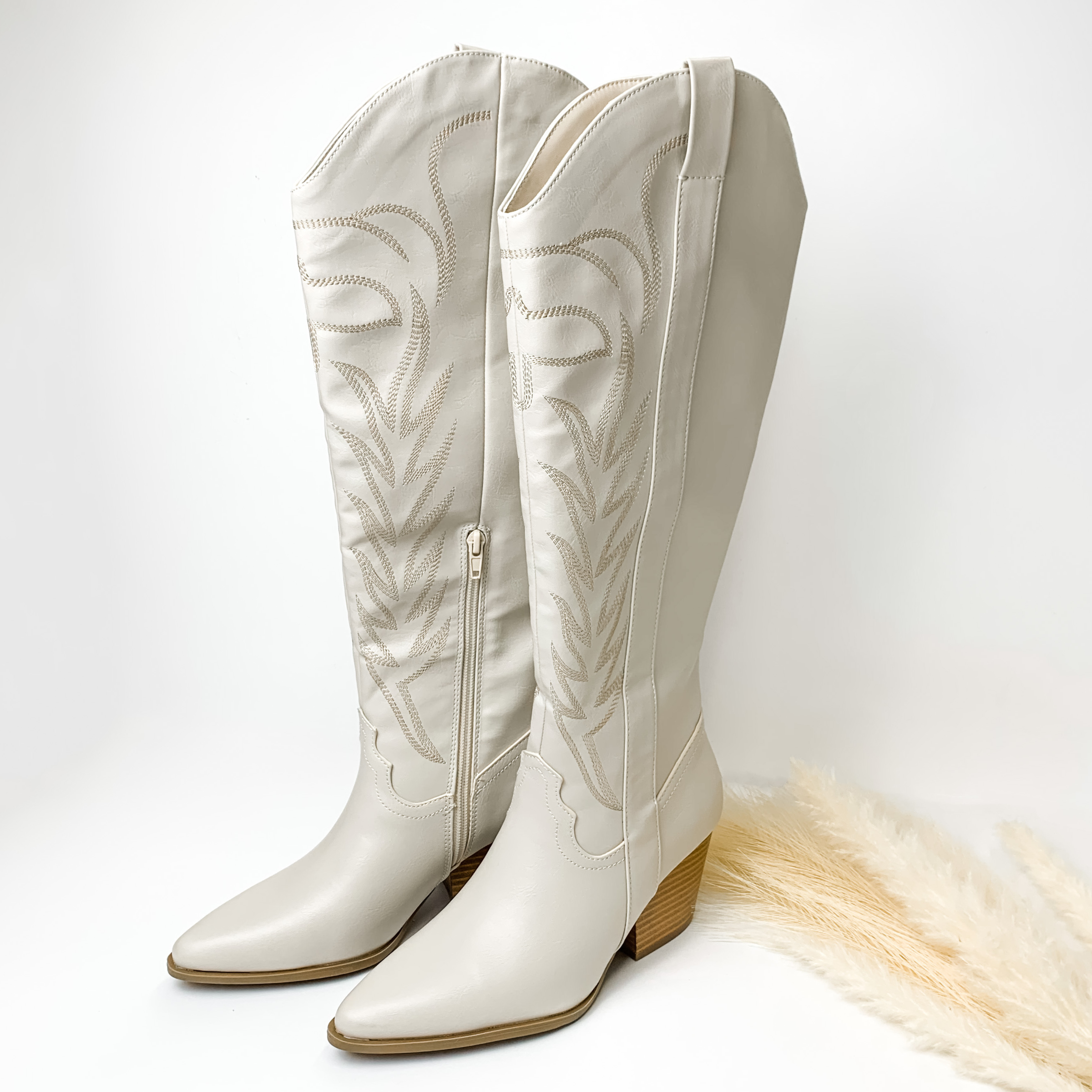 Bone white tall boots with ivory western stitching and tan heel. These boots are pictured on a white background with some ivory pompous grass on the right side of the boots. 