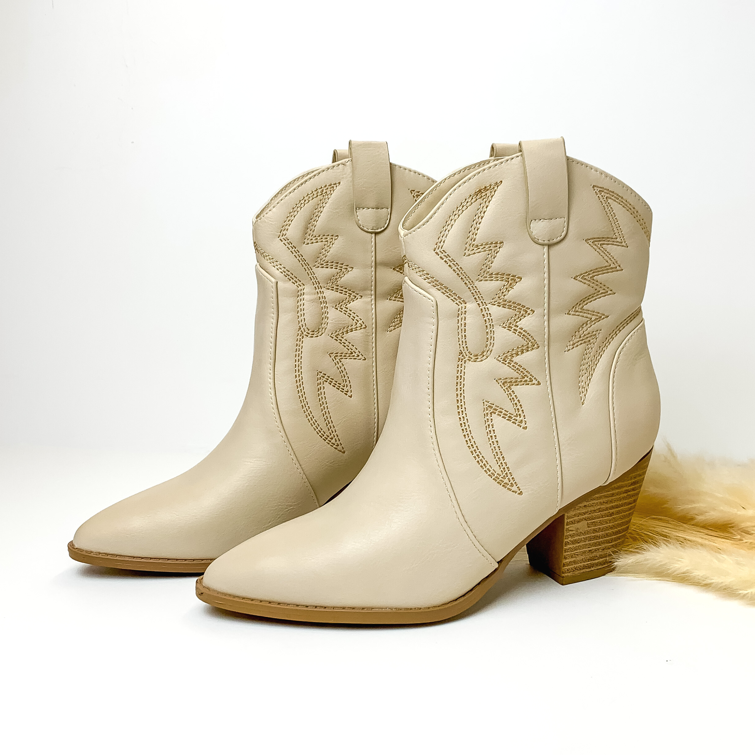Ivory ankle booties with ivory western stitching and black heel. These boots are pictured on a white background with some ivory pompous grass on the right side of the boots. 