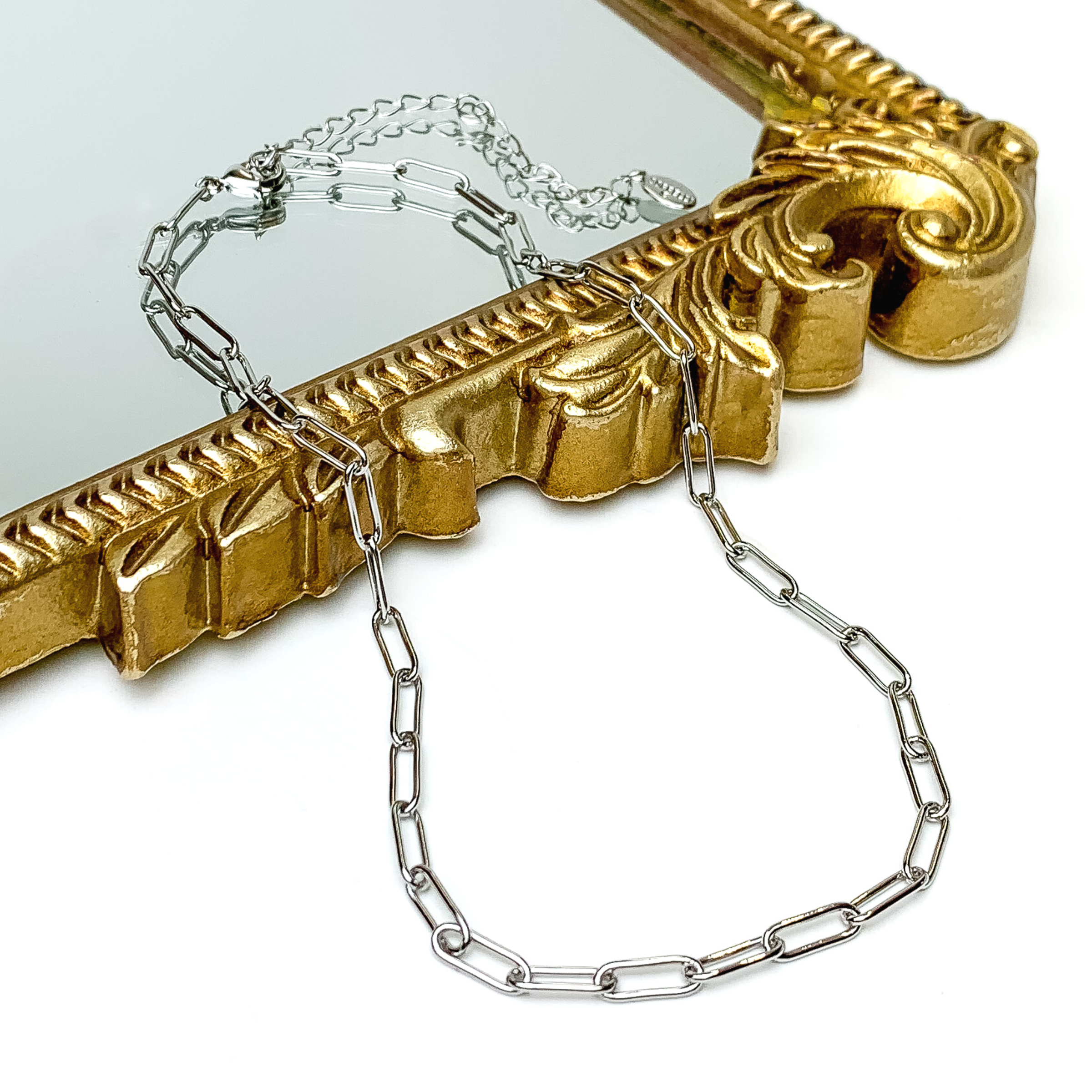 Silver, paperclip chain necklace pictured laying partially on a gold mirror on a white background. 