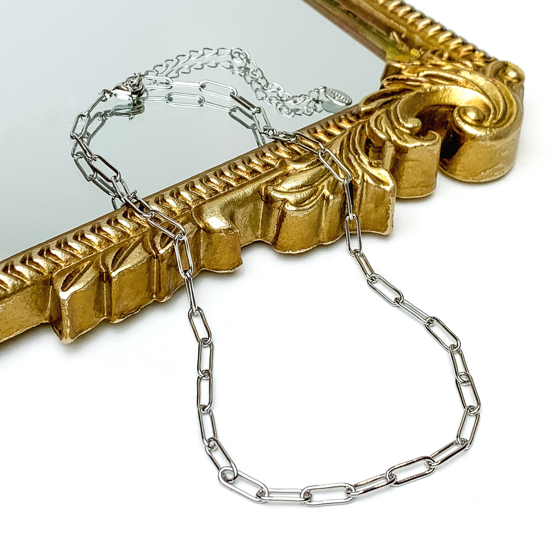 Silver, paperclip chain necklace pictured laying partially on a gold mirror on a white background. 