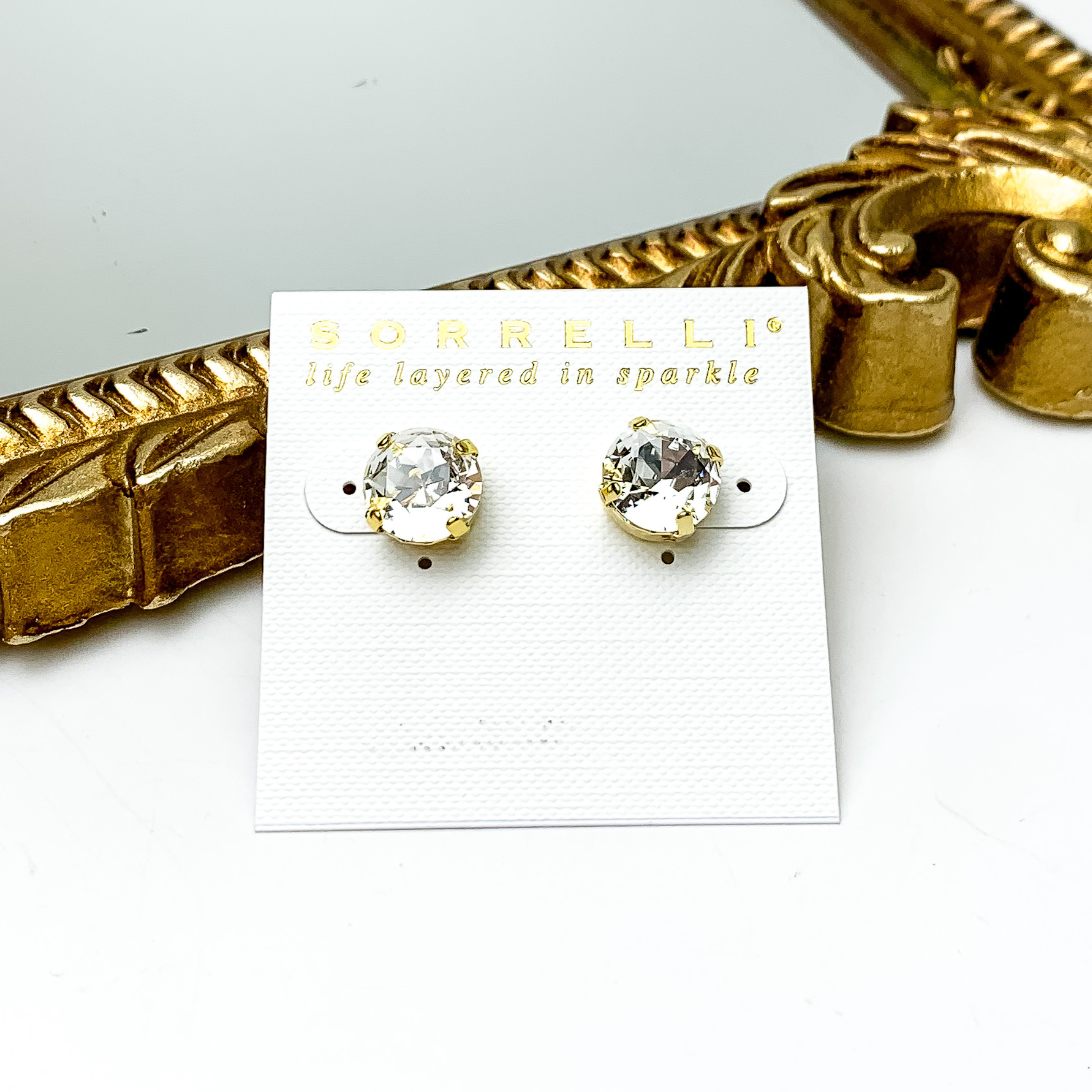 Round, clear crystal stud earrings with gold brackets. These studs are pictured on a white earrings holder in front of a gold mirror on a white background. 
