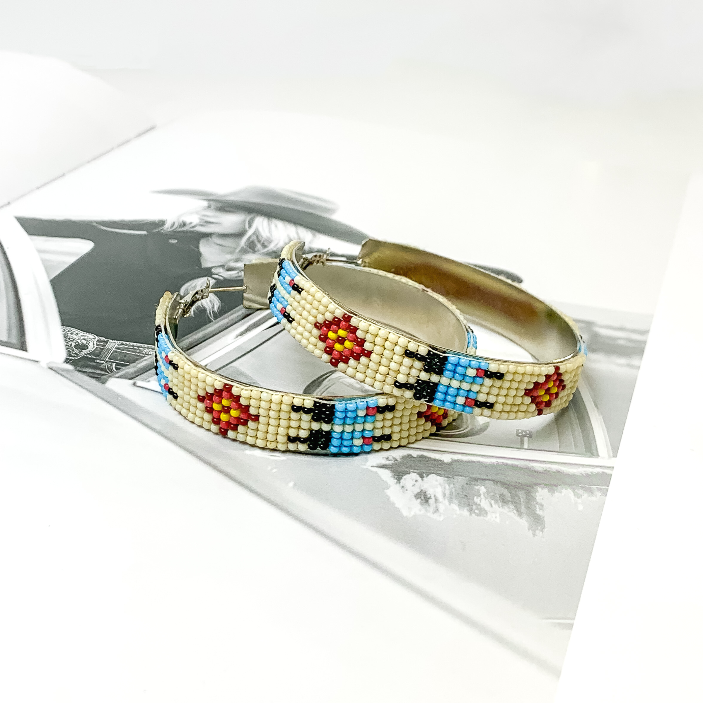 Ivory beaded hoop earrings with a native beaded print. This print includes, white, blue, black, red, and yellow beads. These hoops are pictured on a black and white picture book. 