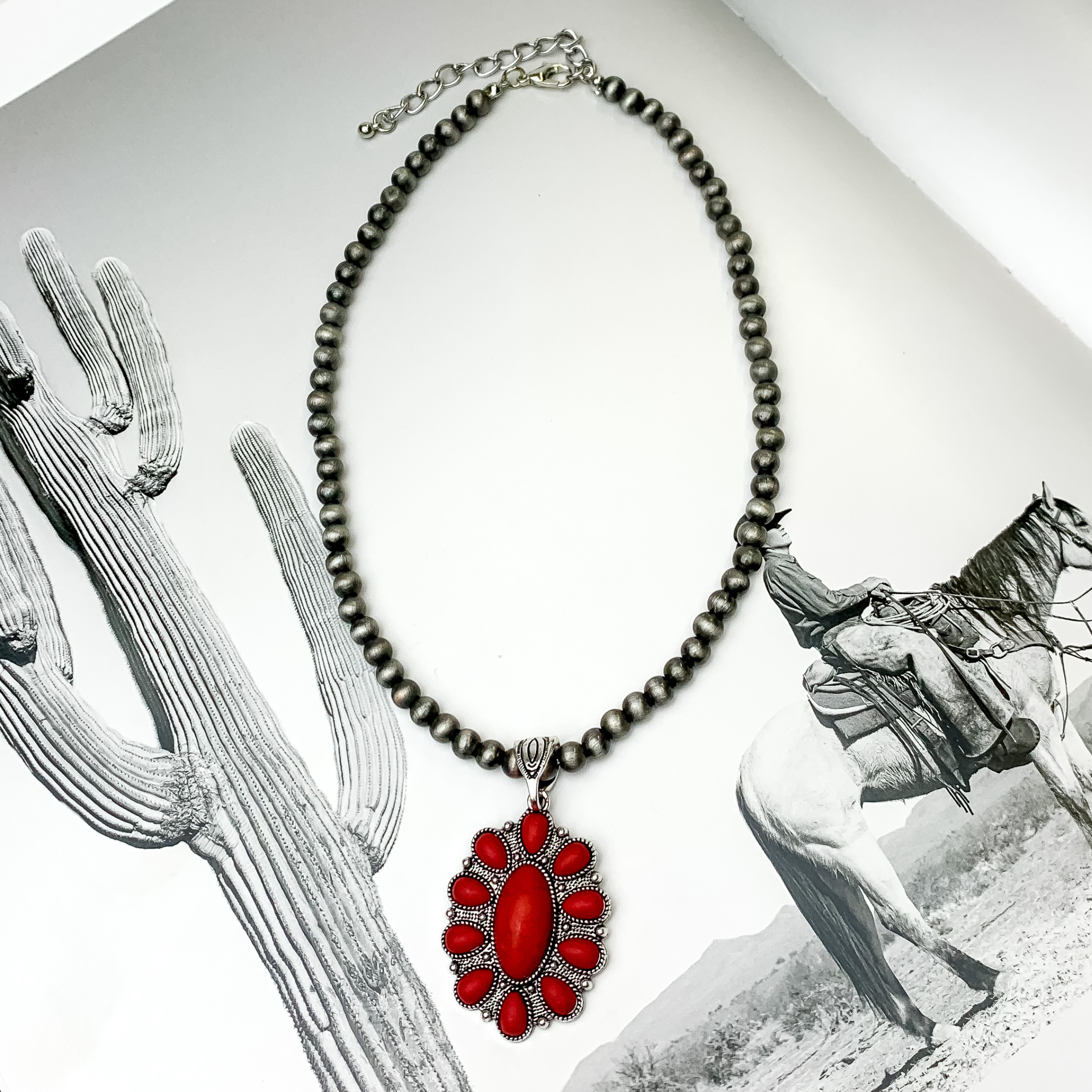 Silver beaded necklace with an oval pendant. This pendant is silver with a red stone design. This necklace is pictured on a black and white picture. 