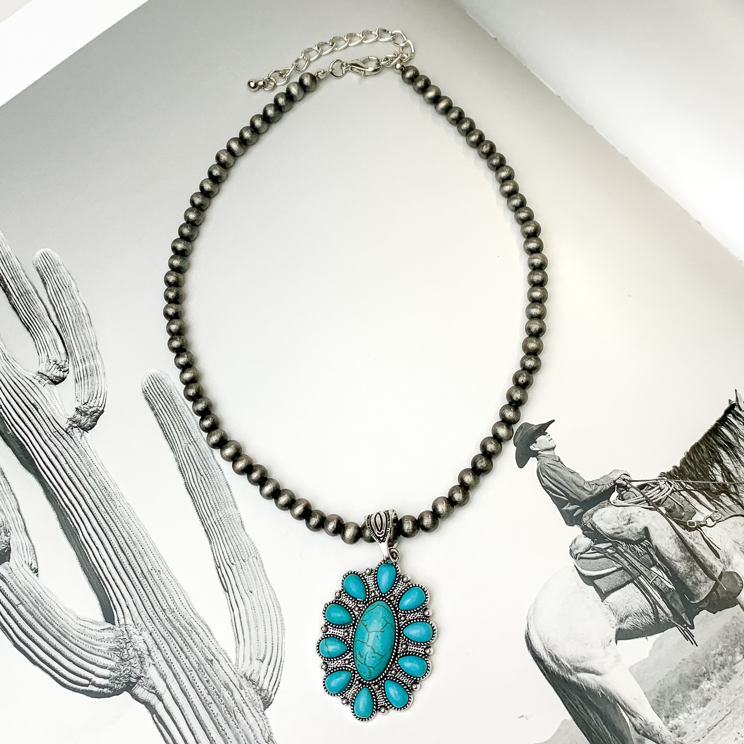 Silver beaded necklace with an oval pendant. This pendant is silver with a turquoise stone design. This necklace is pictured on a black and white picture. 