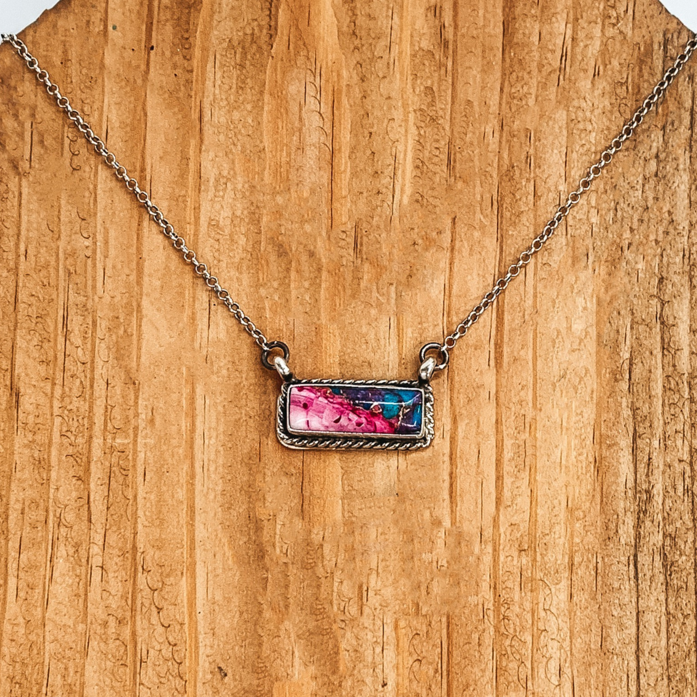 Silver chain necklace with rectangle bar pendant. The pendant has a silver outline with a pink, turquoise, and purple mix of colors. This necklace is pictured on a brown wood background. 