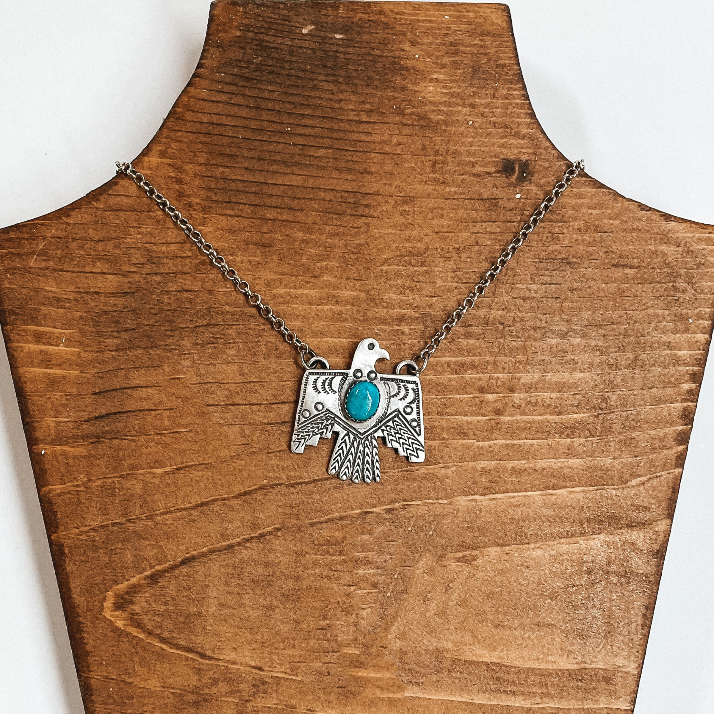 Silver chain necklace with a silver, thunderbird pendant. The thunderbird pendant has a center turquoise stone. This necklace is pictured on a wood necklace holder on a white background. 