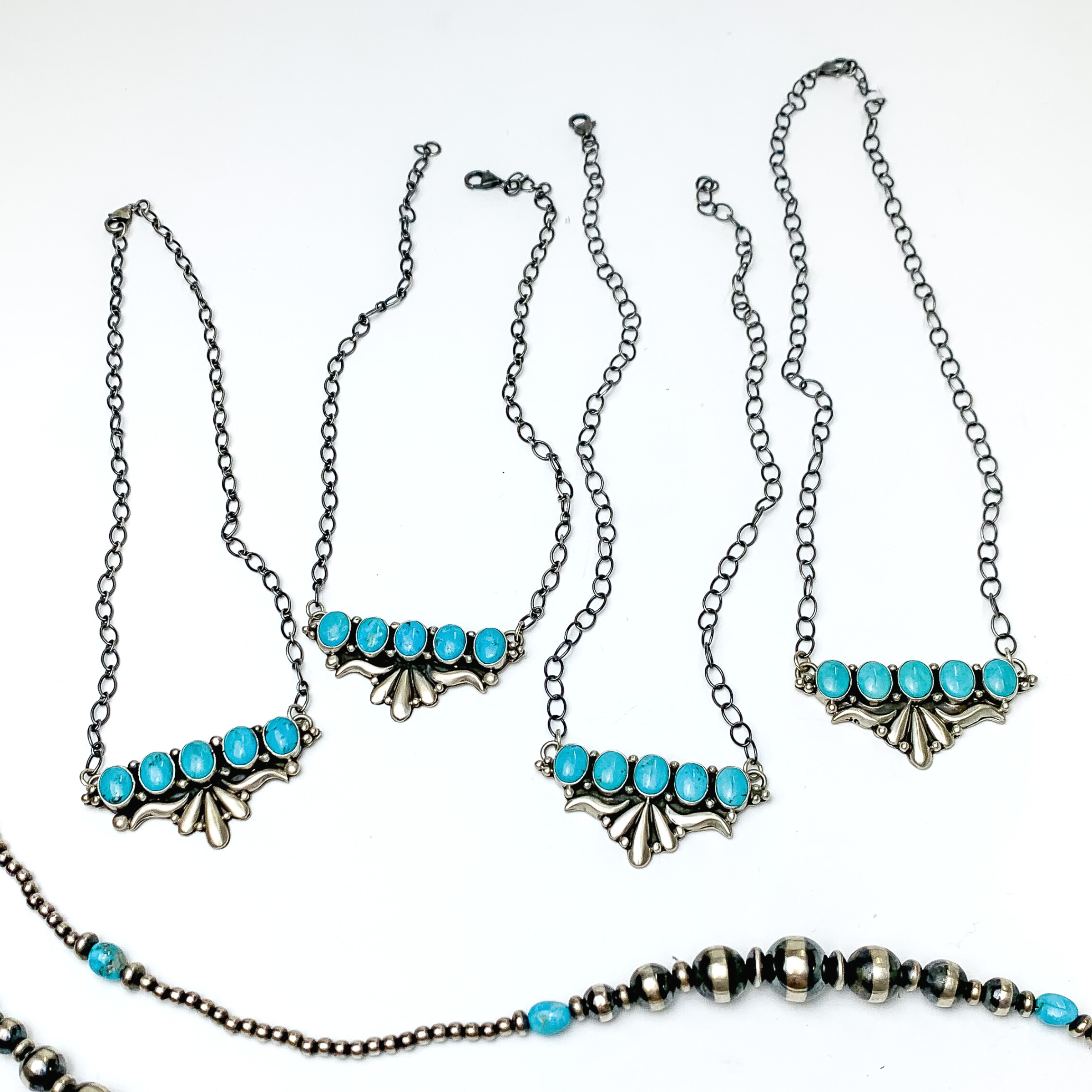 Four silver chain necklace with a silver and turquoise stone pendant. The pendant has some silver detailing and five turquoise stones. These necklaces are pictured on a white background with silver beads at the bottom of the picture.  