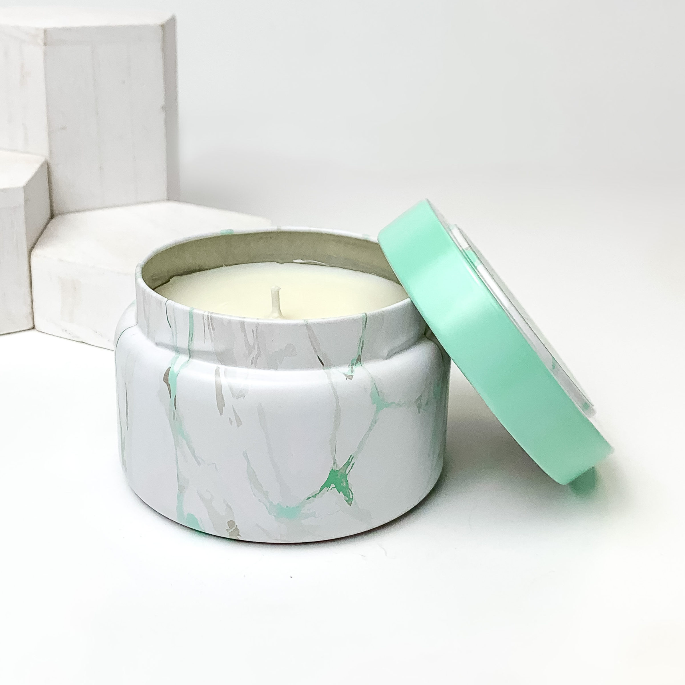 White, grey and mint marble candle with the lid leaning on the side of the candle. This candle is pictured in front of white blocks on a white background. 