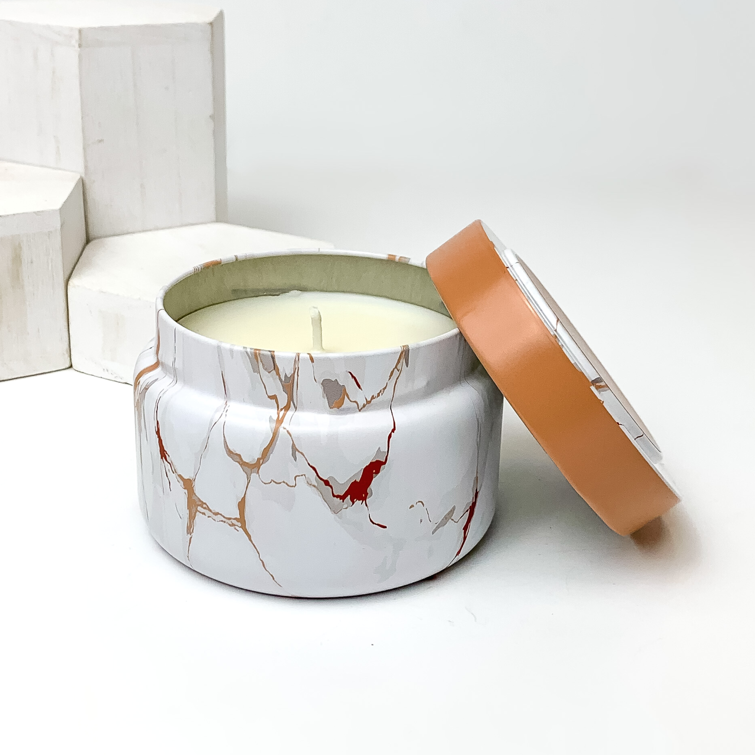 White, grey and red/brown marble candle with the lid leaning on the side of the candle. This candle is pictured in front of white blocks on a white background. 