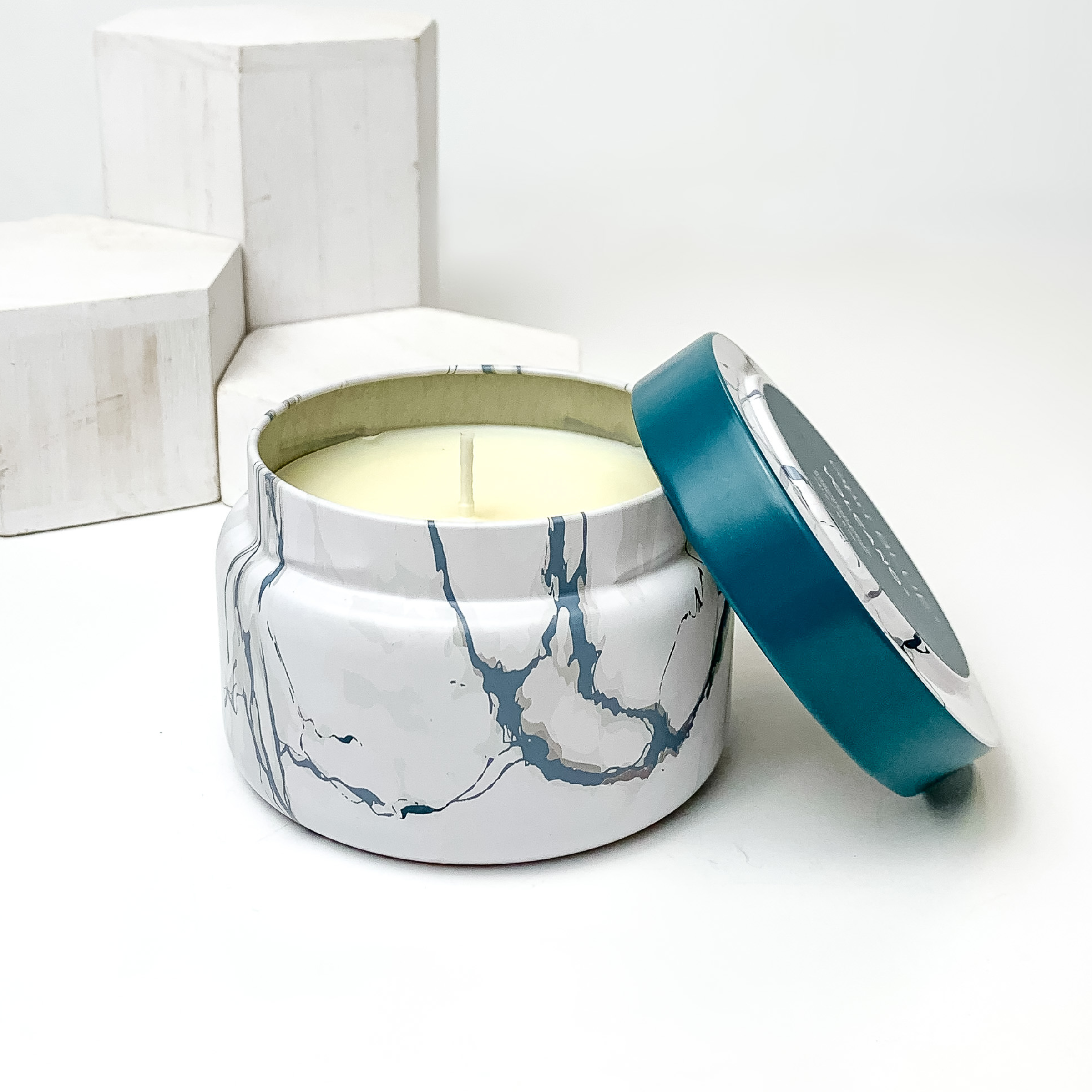 White, grey and dark blue marble candle with the lid leaning on the side of the candle. This candle is pictured in front of white blocks on a white background. 