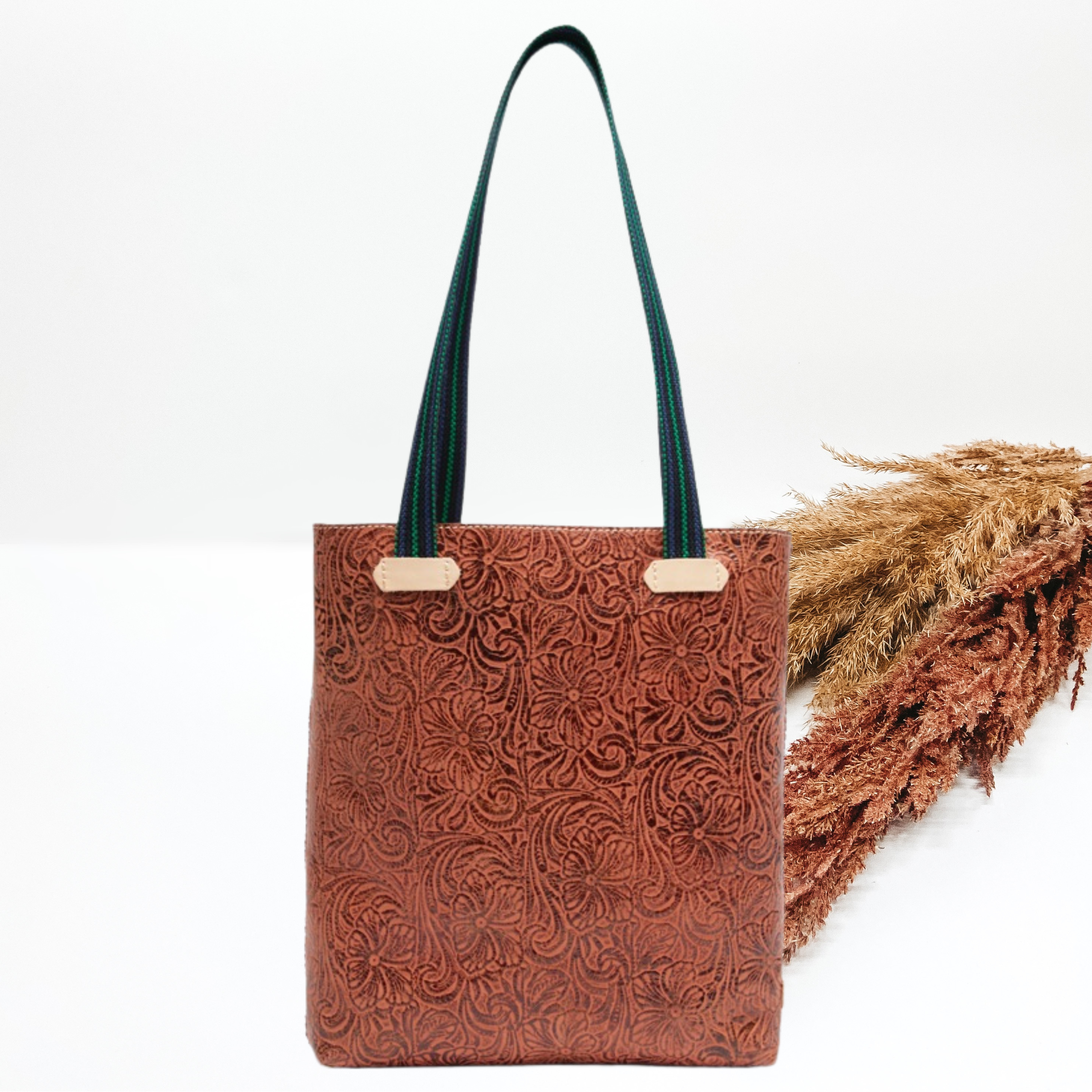Pictured is long, rectangle tote that has a leather tooled all over print. This purse also includes striped purse straps. This purse is pictured on a white background with pompous grass on the right side of the picture.