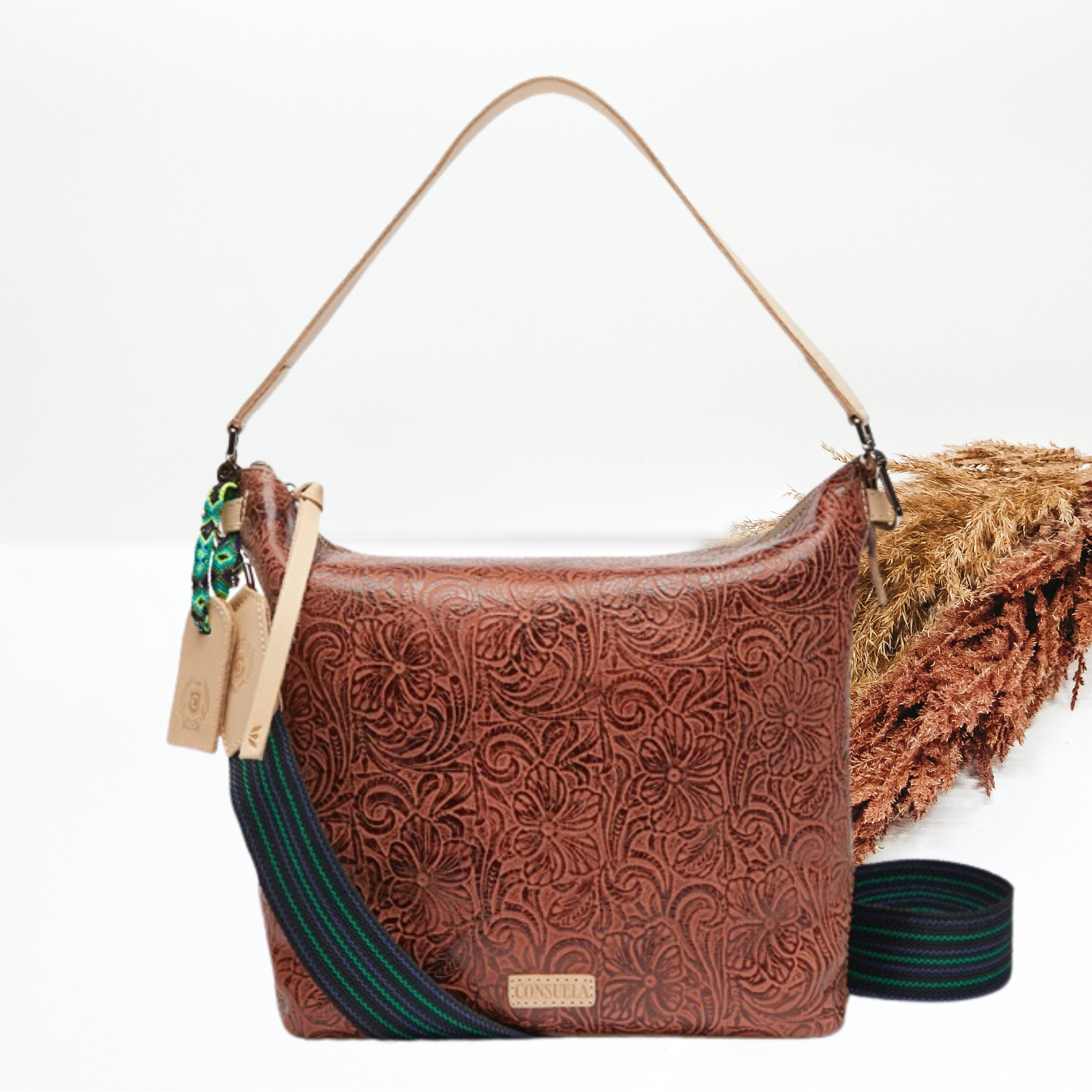 Pictured is hobo purse that has a tooled leather all over deisgn. This purse includes a thick, light tan strap and striped, thick purse strap. This purse is pictured on a white background with pompous grass on the right side of the picture.