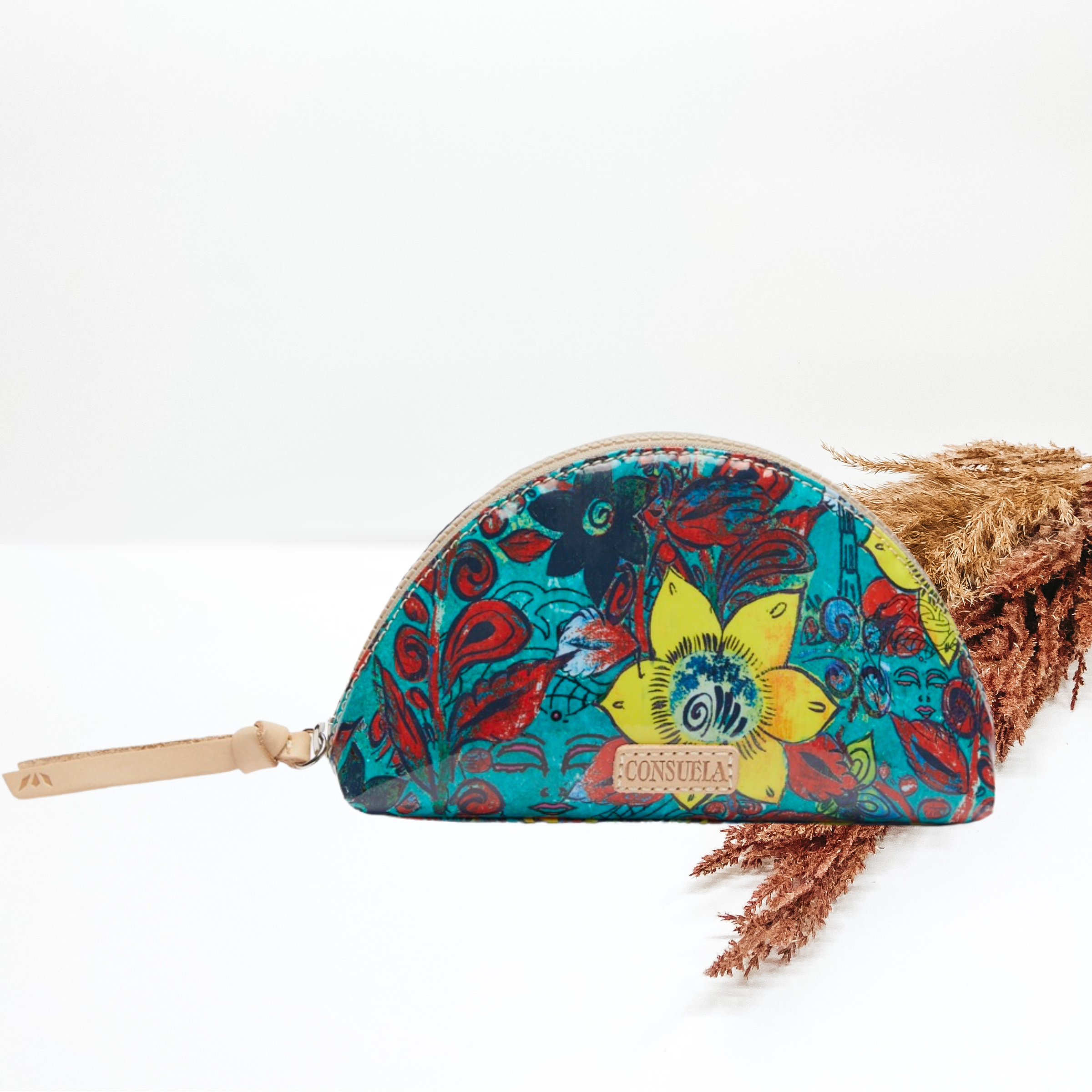 Pictured is a dome shaped cosmetic bag. This is a blue bag with a navy, yellow, and red print design. It also includes a top zipper. This purse is pictured on a white background with pompous grass on the right side of the picture.