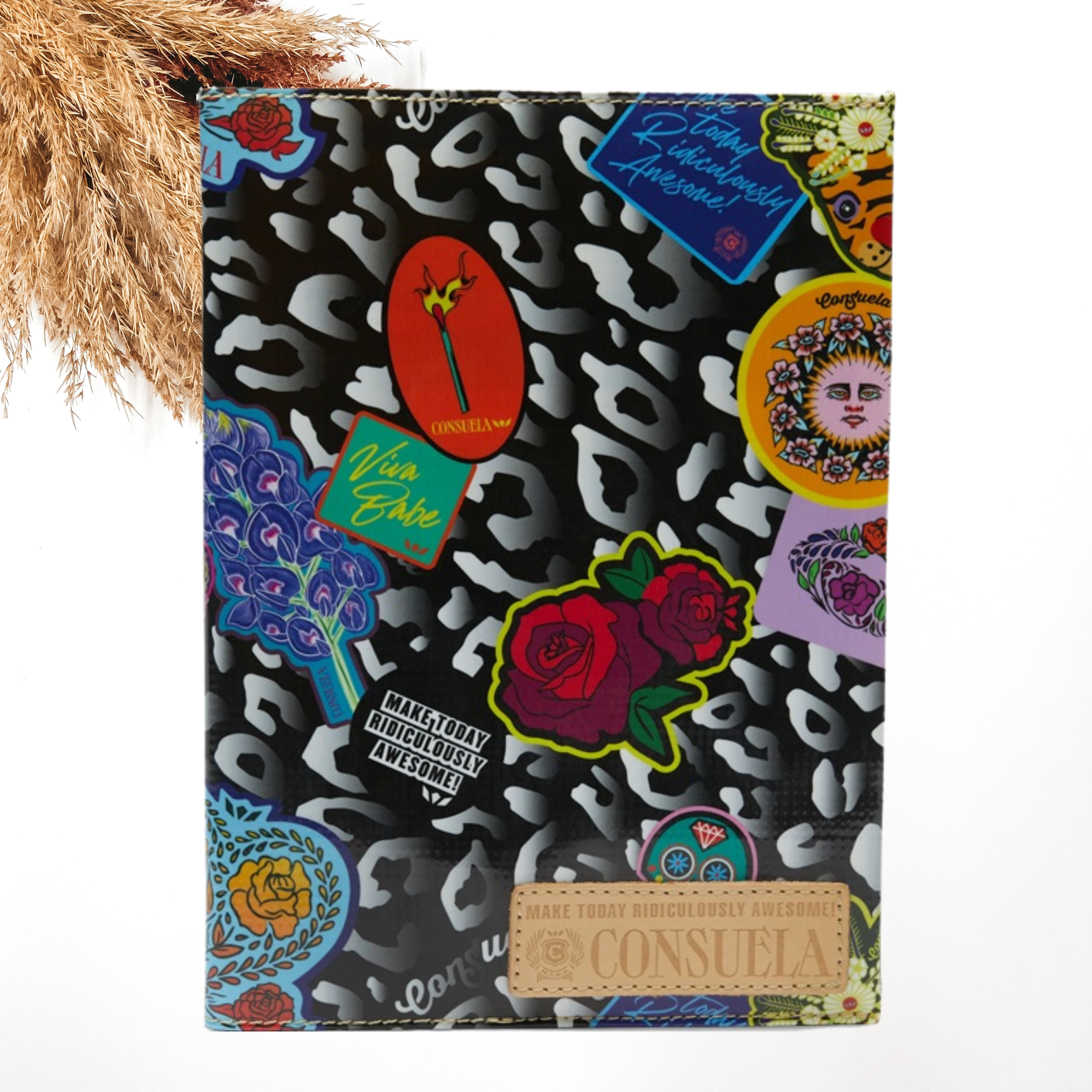 Pictured is a notebook cover with a mostly black with grey leopard print design. It also includes random sticker patches throughout and a light tan Concsuela patch on the bottom right corner. This notebook is pictured on a white background with pompous grass in the top left corner of the picture.